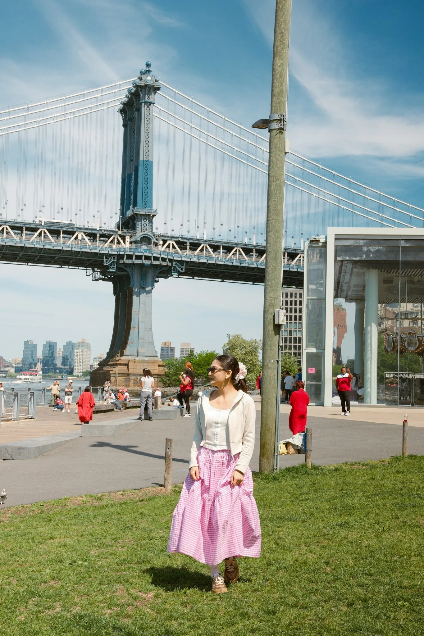 A woman wearing a white blouse and pink checkered skirt walking on green grass near a waterfront with a bridge and cityscape in the background.