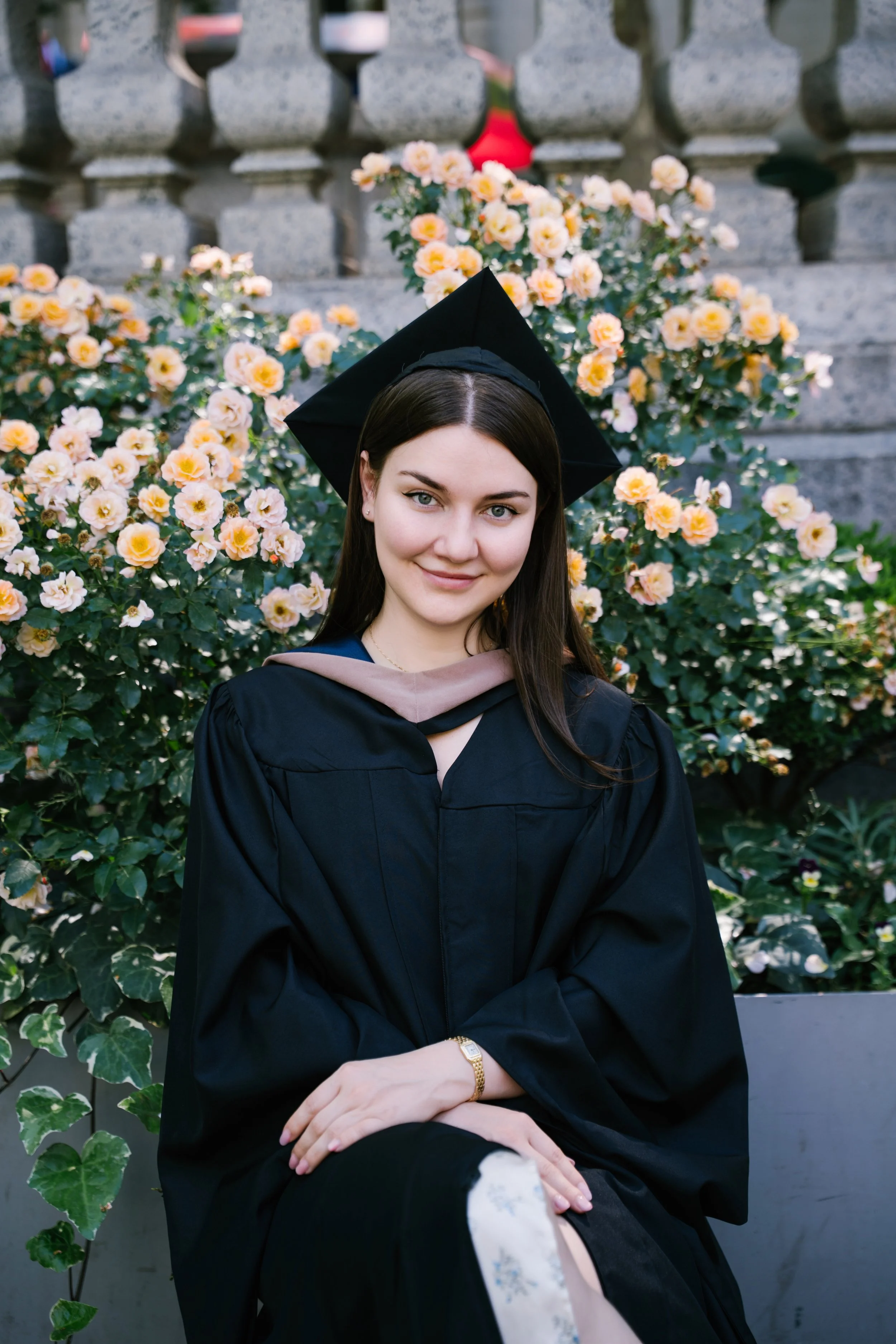 A young woman in a graduation cap and gown sitting in front of a stone wall with blooming pink and yellow roses, smiling at the camera.