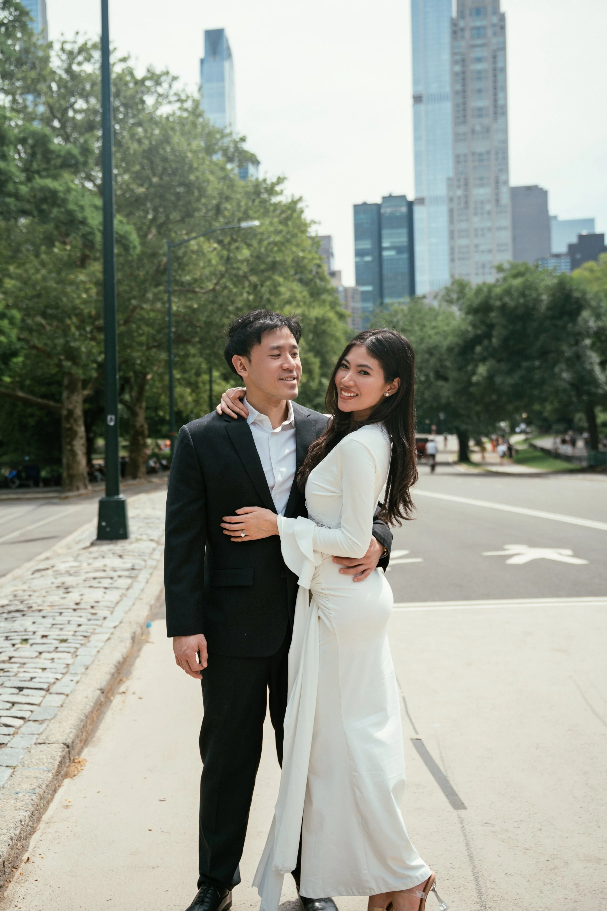 A couple dressed in formal attire is standing together outdoors with a cityscape in the background, including tall buildings and green trees.