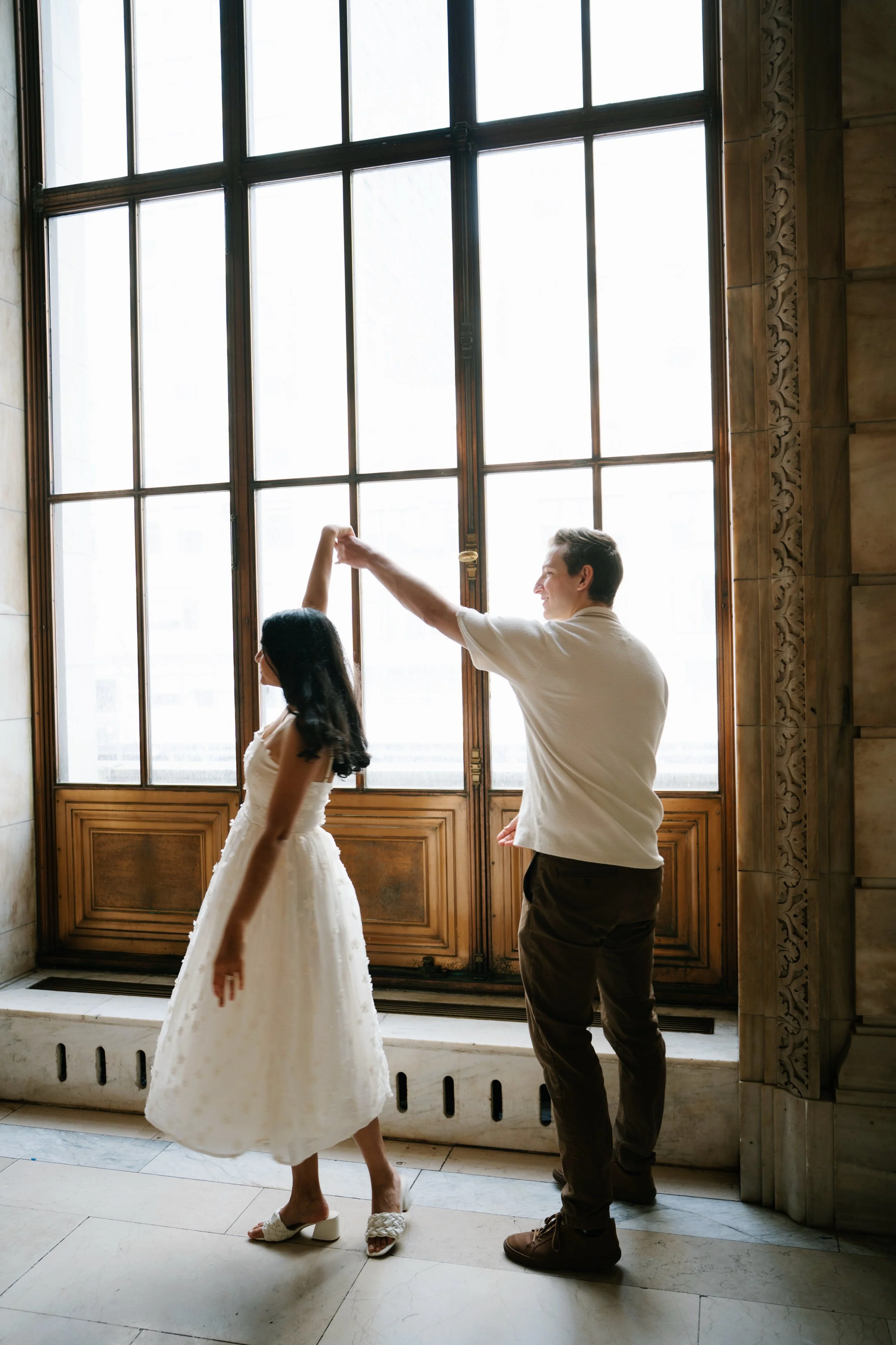 A man and a woman dancing in front of large window with decorative wood trim. The woman is wearing a white dress and the man is wearing a white shirt and dark pants.