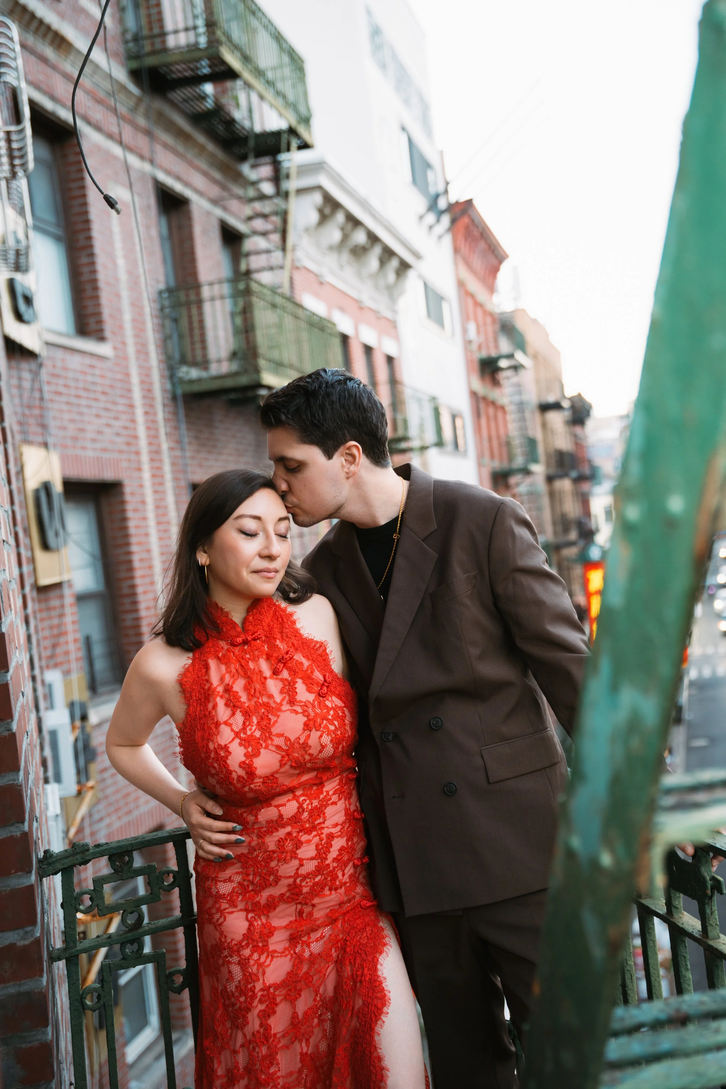 A young man with dark hair kissing a young woman on the forehead, dressed in a red lace dress, on a city balcony with brick buildings in the background.