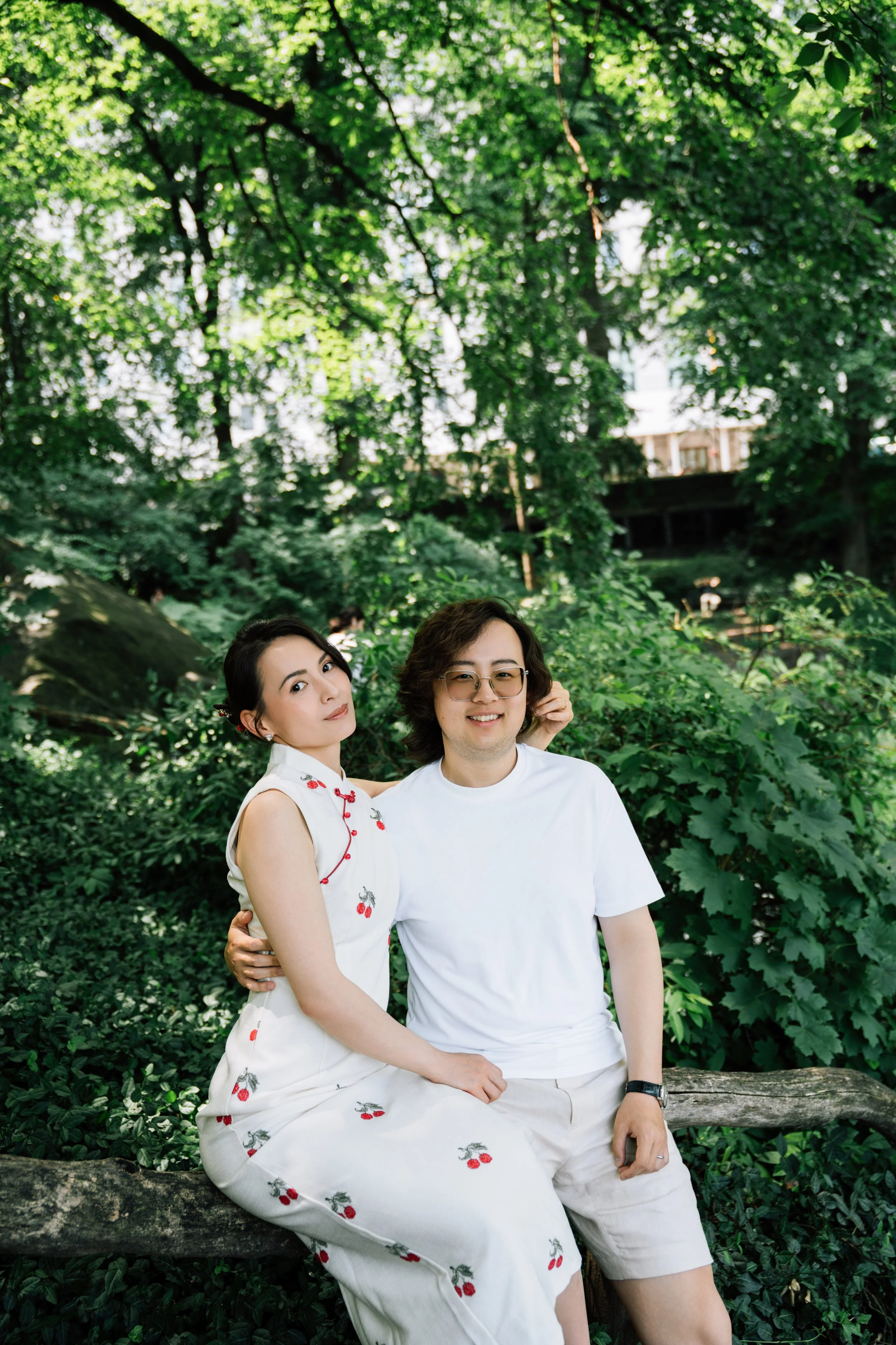 A woman and a man sitting on a log in a lush green forest, with trees and dense foliage behind them. The woman is wearing a white dress with cherry patterns, and the man is wearing a white t-shirt and light-colored shorts.