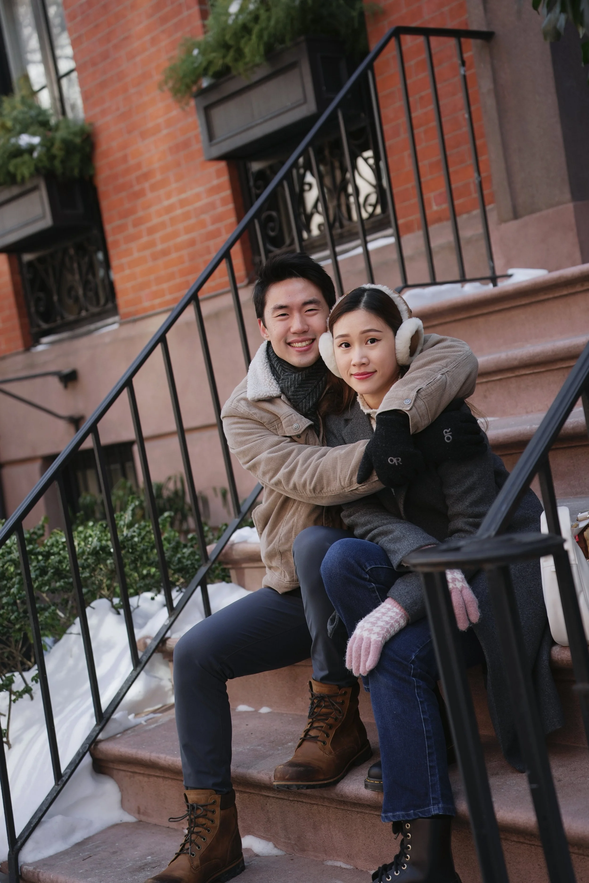 A young couple sitting on the steps of a brownstone building in winter. The man is smiling, with his arms around the woman, who is wearing earmuffs and gloves. Snow is visible on the stairs and in the garden below.