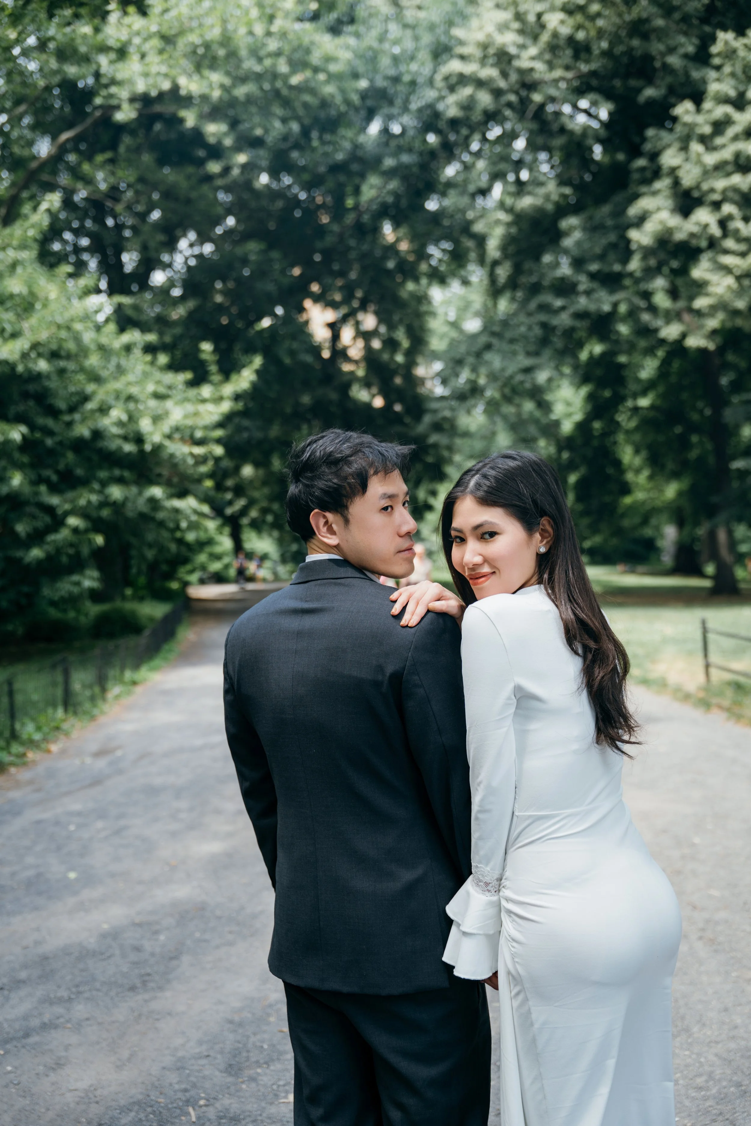 A couple, dressed in formal attire, standing on a park pathway with lush green trees around them. The woman, with long dark hair, is wearing a white dress and the man, with short dark hair, is wearing a dark suit. They are looking at the camera. The 