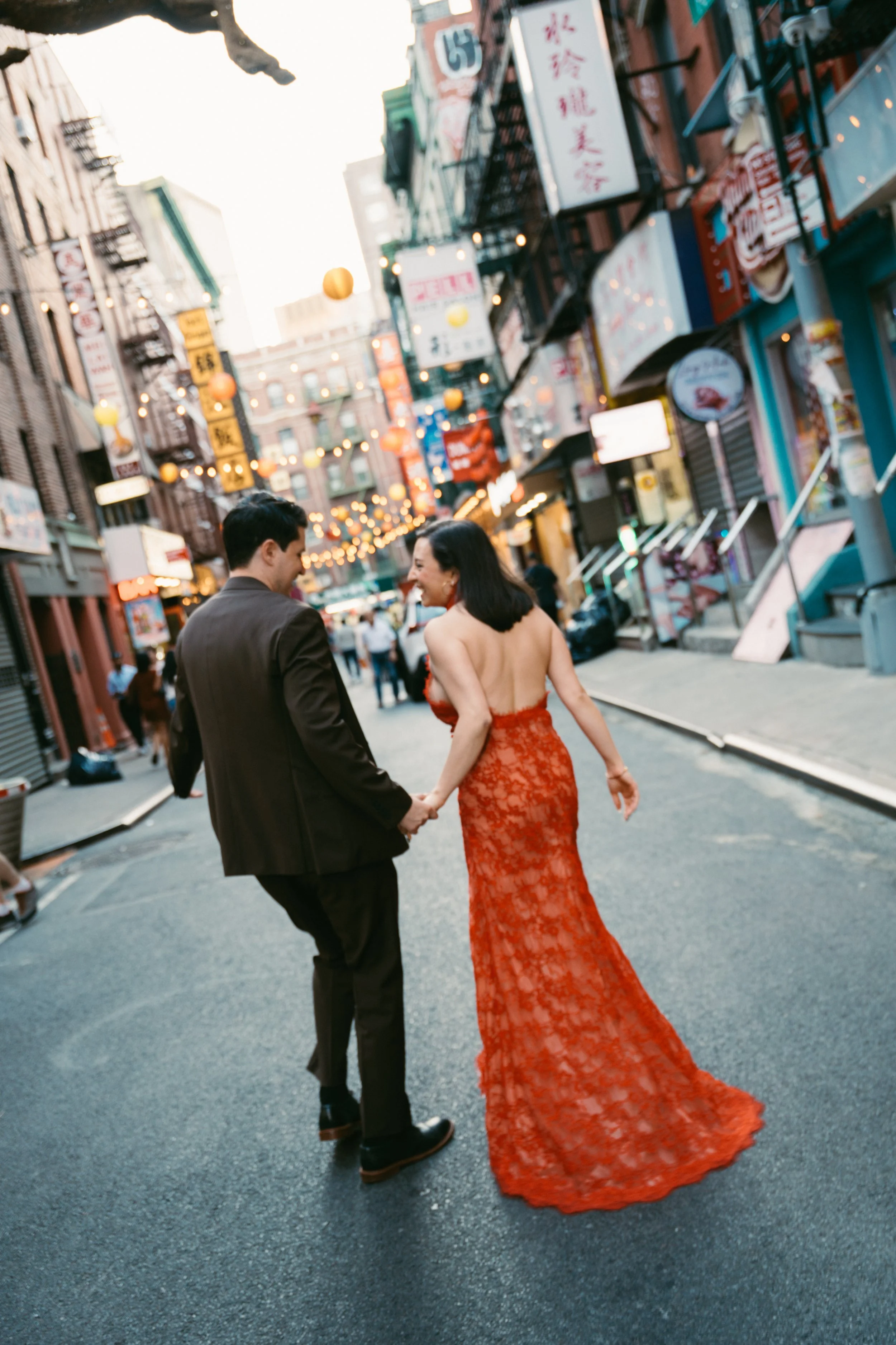 A man and a woman holding hands and walking on a city street decorated with hanging lanterns and signs in an Asian city at sunset, with the woman dressed in a red lace gown.