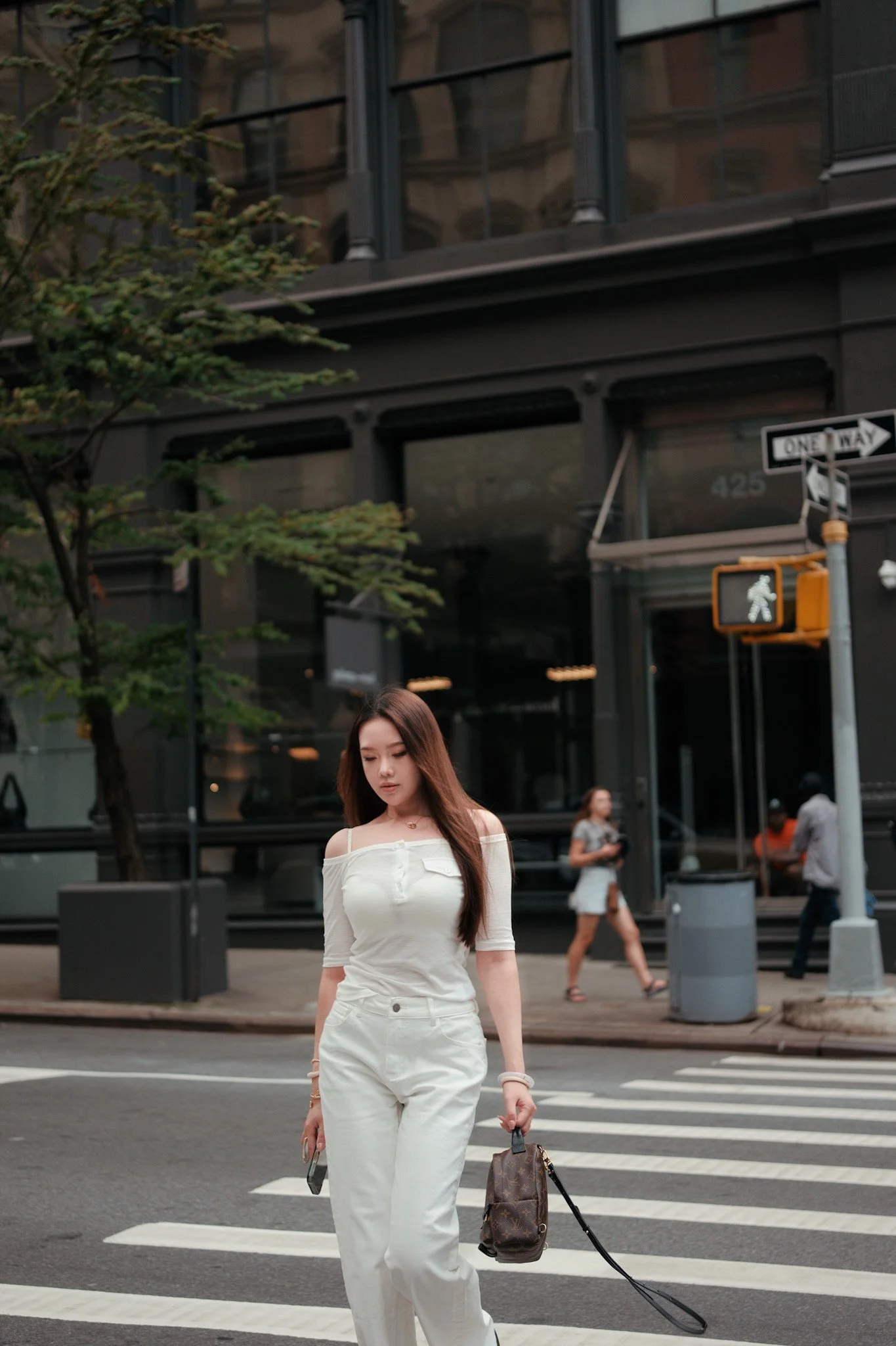 Young woman with long brown hair walking across a crosswalk in an urban setting, carrying a handbag and a phone, with a glass building and other pedestrians in the background.