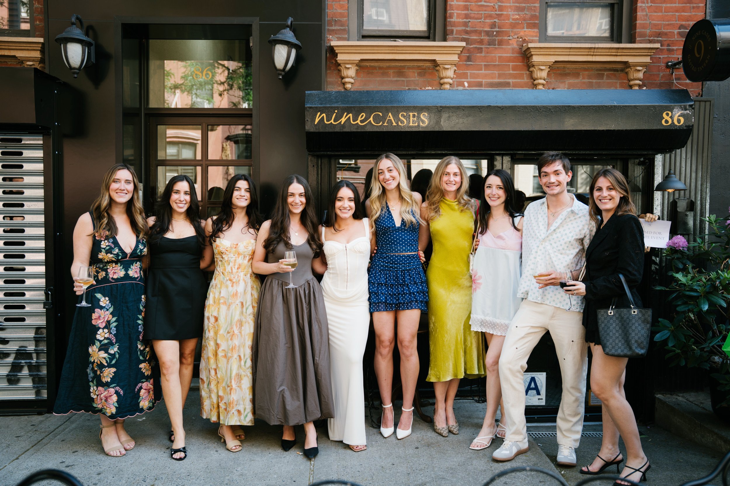 A group of ten young adults, mostly women and one man, standing together outside a restaurant named 'nine CASES' at 86, smiling at the camera, some holding wine glasses, in casual and semi-formal attire.