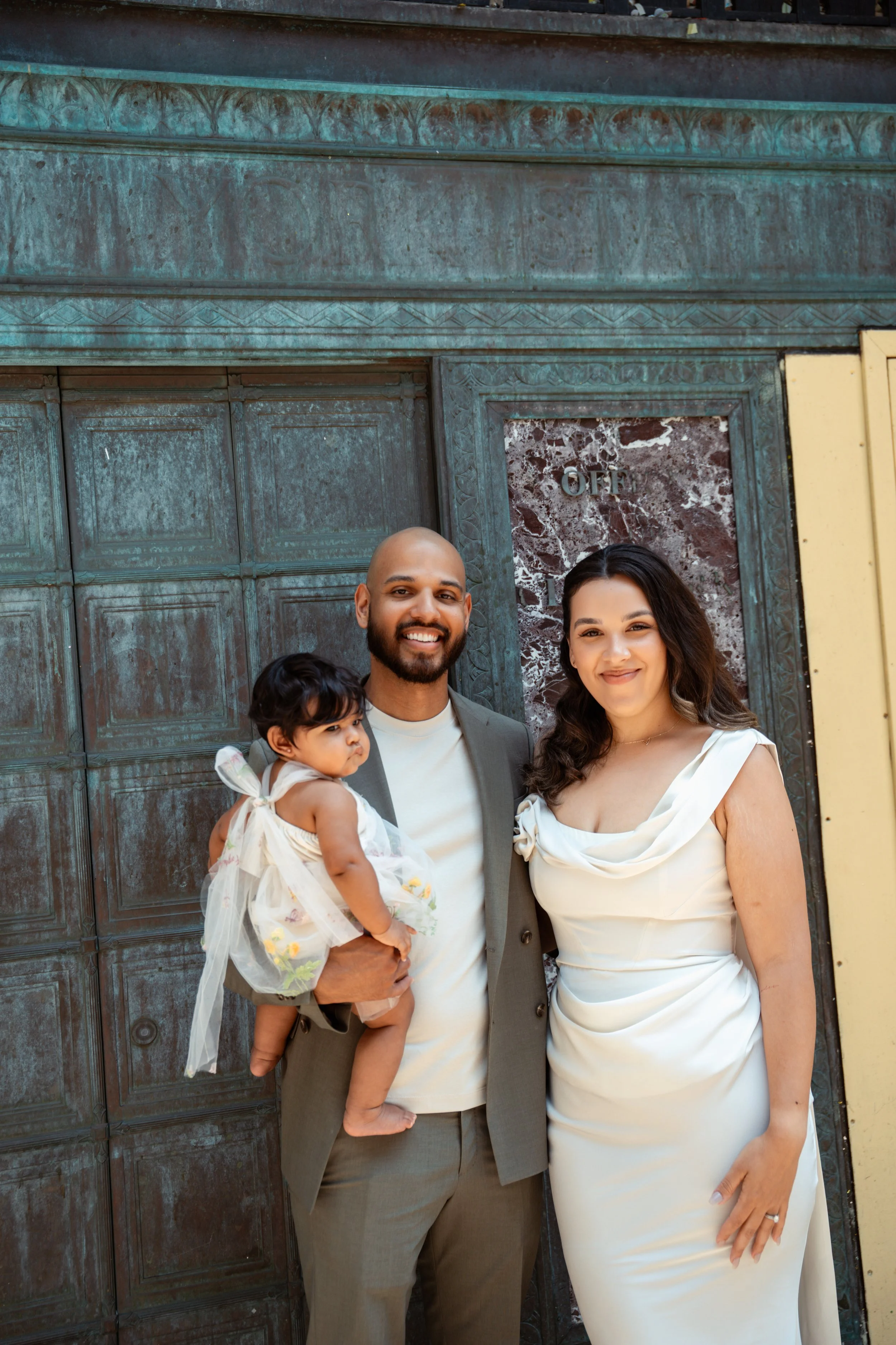 A happy family of three, including a man, woman, and a young girl, standing together outdoors in front of a large, ornate metal door with intricate designs, after a wedding or formal event. The woman wears a white dress, the man wears a gray suit, an
