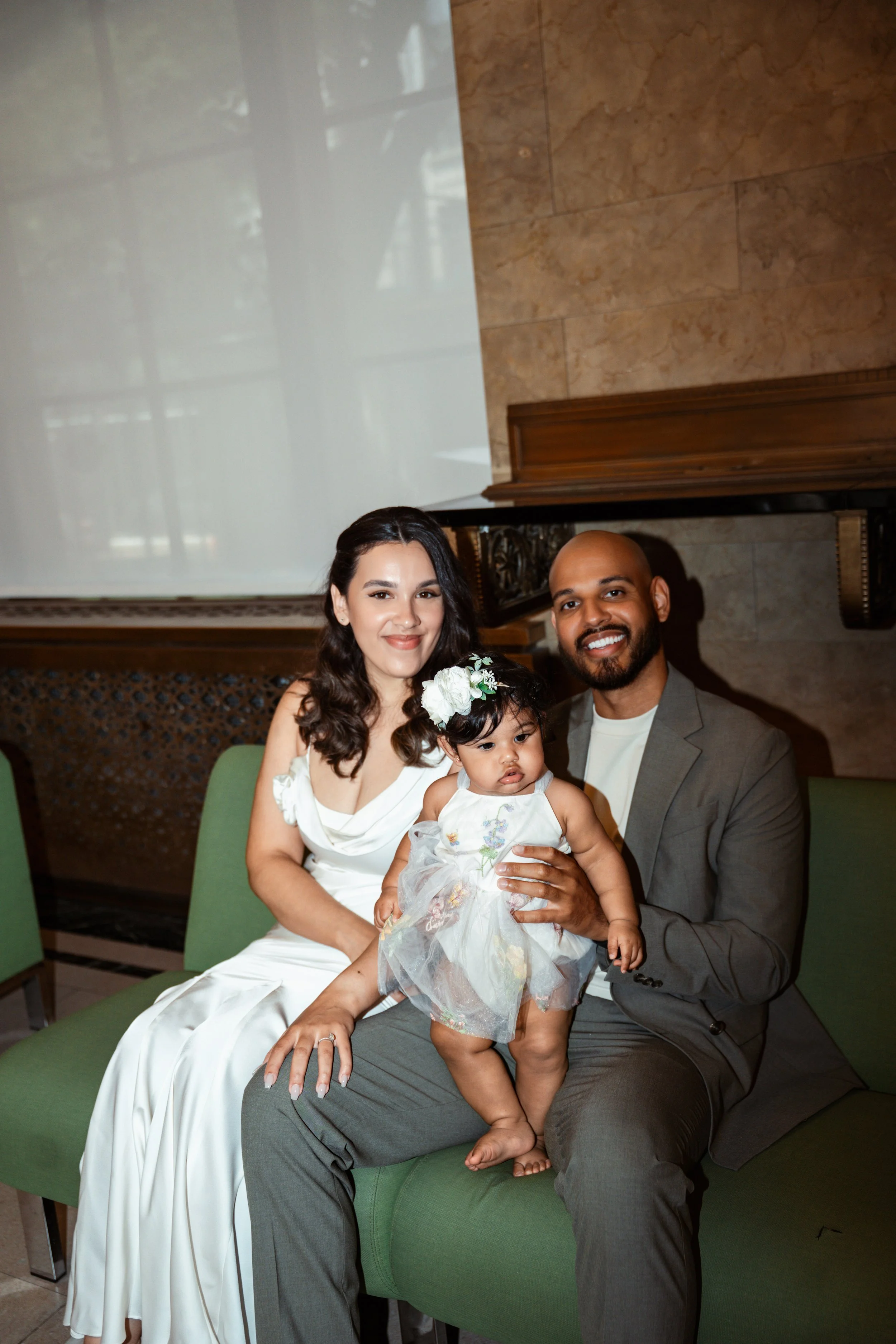 A family of three sitting on a green sofa in a room with a marble wall. The woman is wearing a white dress, holding a young girl dressed in a white dress with floral embroidery, and the man is wearing a gray suit. All three are smiling, and the woman