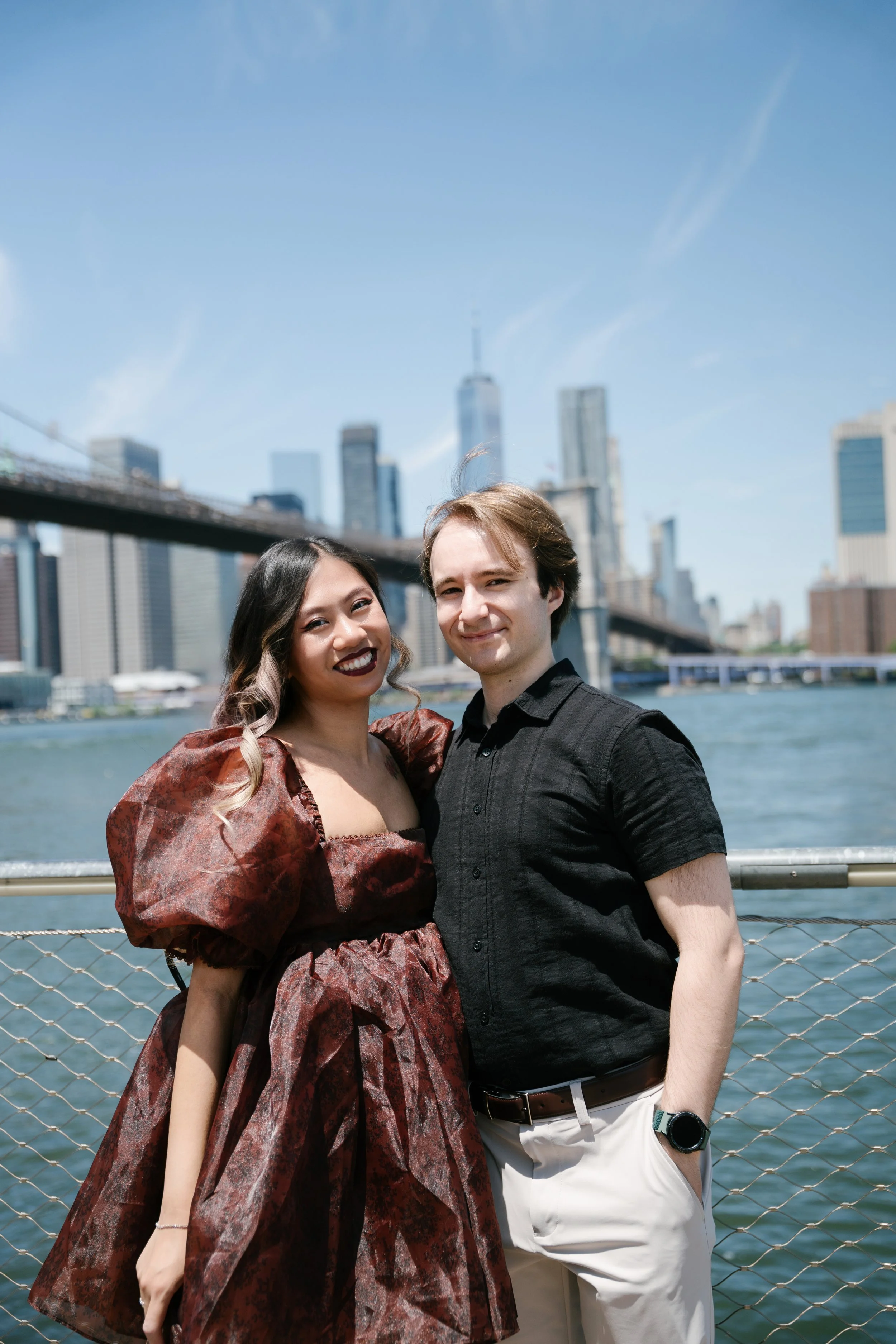A smiling woman with wavy hair wearing a patterned dress and a man with short hair wearing a black shirt, standing by a river with a city skyline and a bridge in the background.