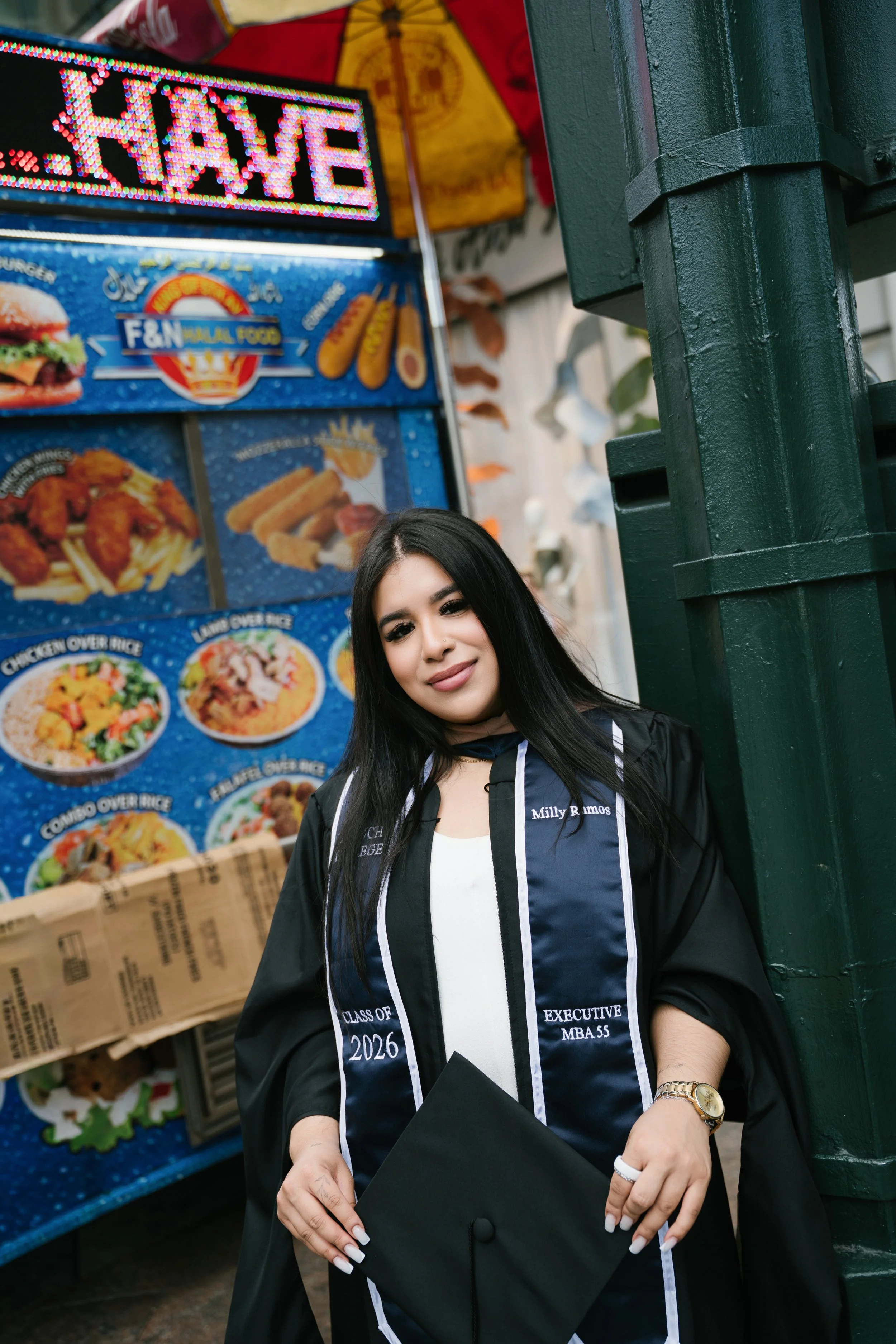 A woman in a graduation gown standing outdoors near a food stand with a menu, holding a black folder or diploma cover, smiling at the camera.