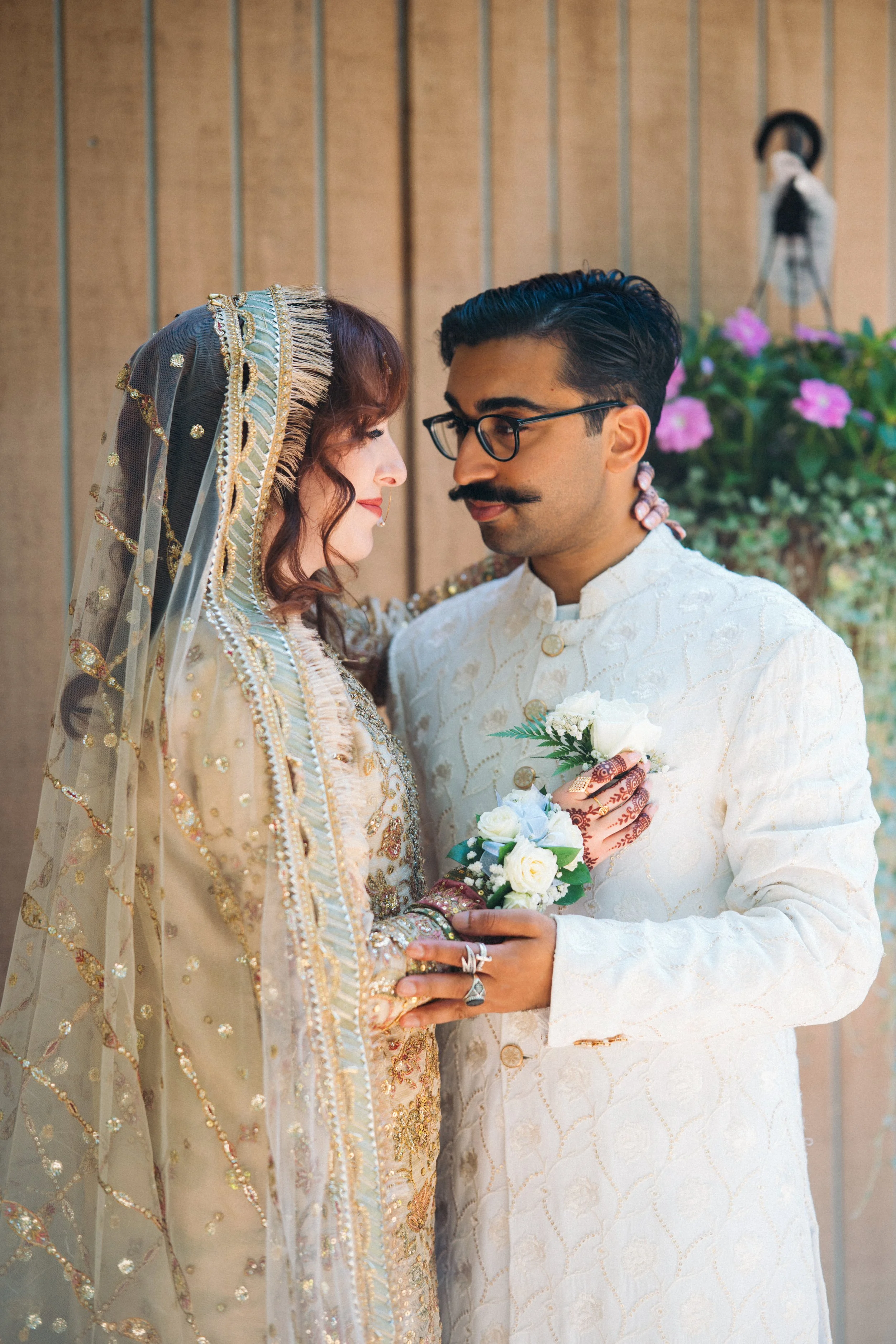A bride and groom dressed in traditional South Asian wedding attire sharing an intimate moment, with the bride wearing a heavily embroidered beige and gold outfit with a matching veil, and the groom in a cream-colored sherwani holding a bouquet of wh