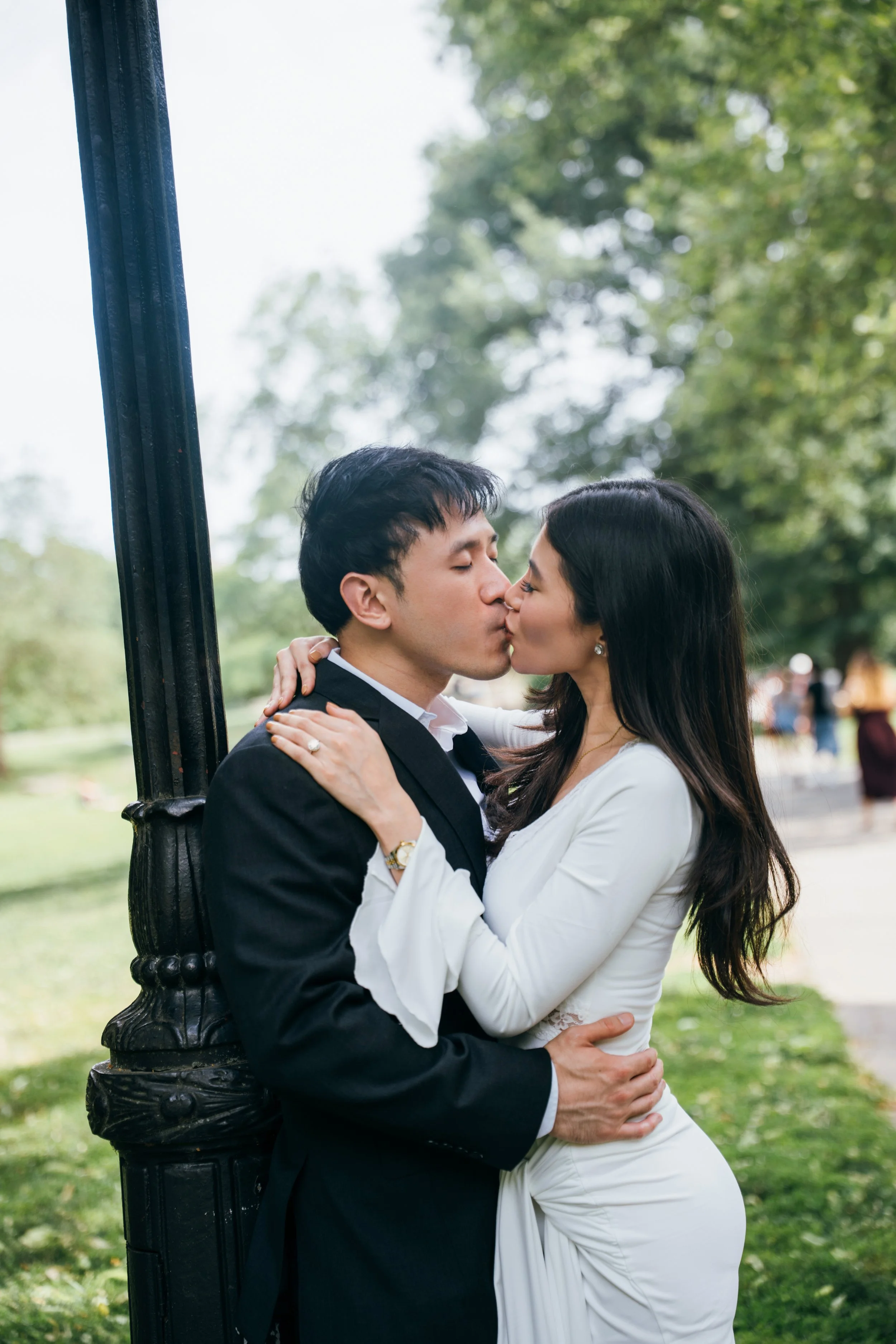 A newlywed couple passionately kissing outdoors, standing close to a black lamppost in a park with green trees in the background.