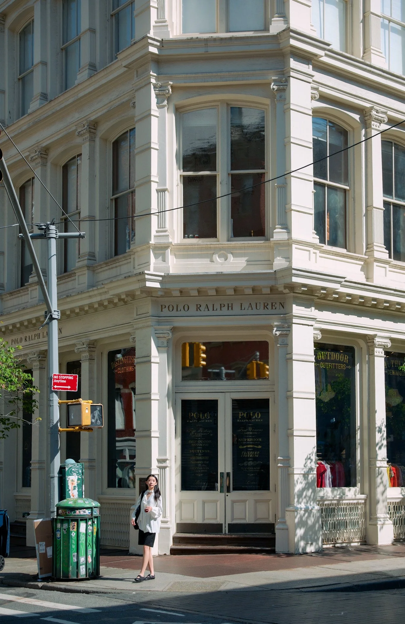 Corner of a white historic building with large windows and a sign reading "Polo Ralph Lauren." A woman in a white top, black skirt, and sandals stands on the sidewalk in front of the building, holding a coffee cup. A person is partially visible insid