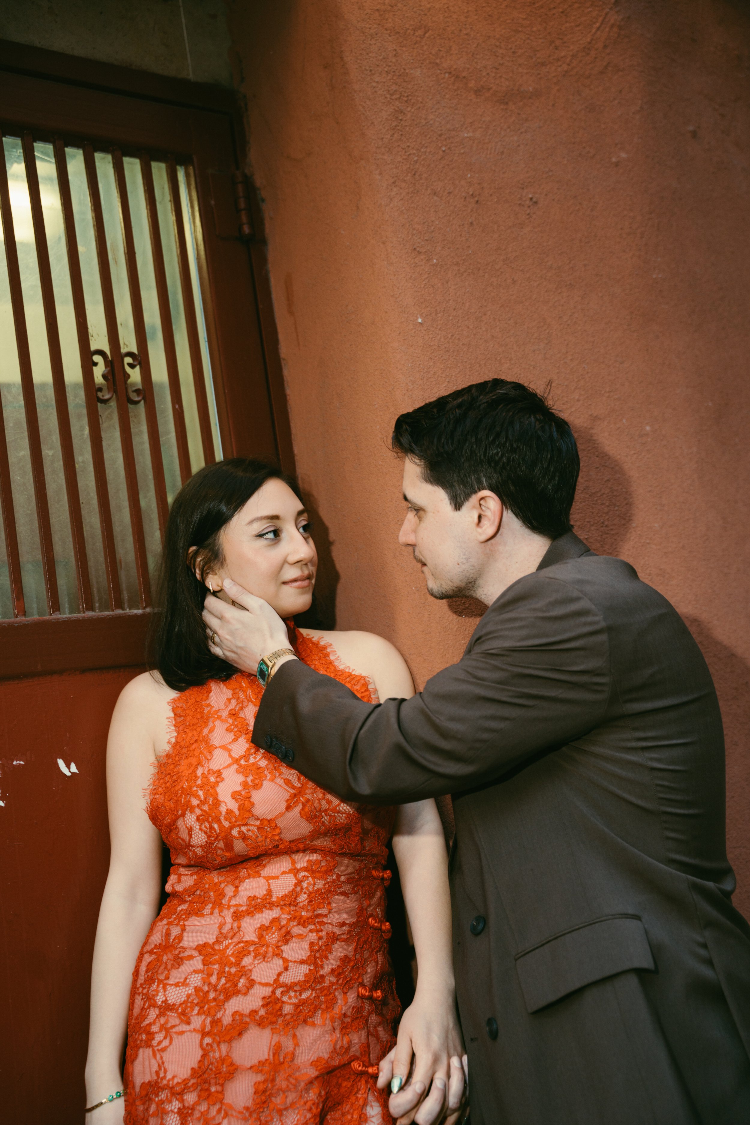 A man and a woman are sitting closely against a red wall. The woman is wearing an orange lace dress and has her hand gently touching her neck. The man is wearing a dark suit and is holding her hand while gazing at her.