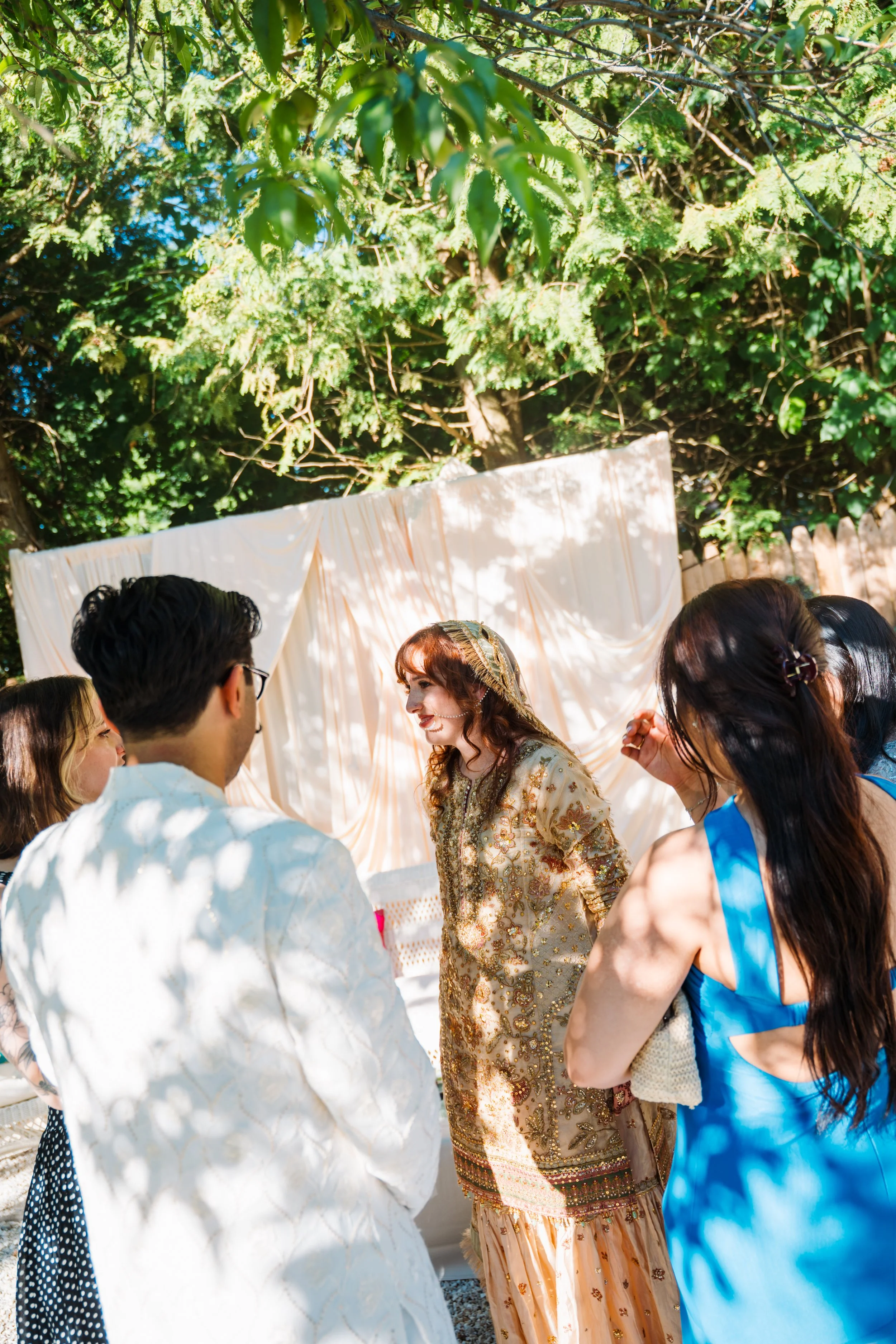 Group of people dressed in traditional South Asian attire participating in a wedding ceremony outdoors, with greenery and a white draped fabric background.