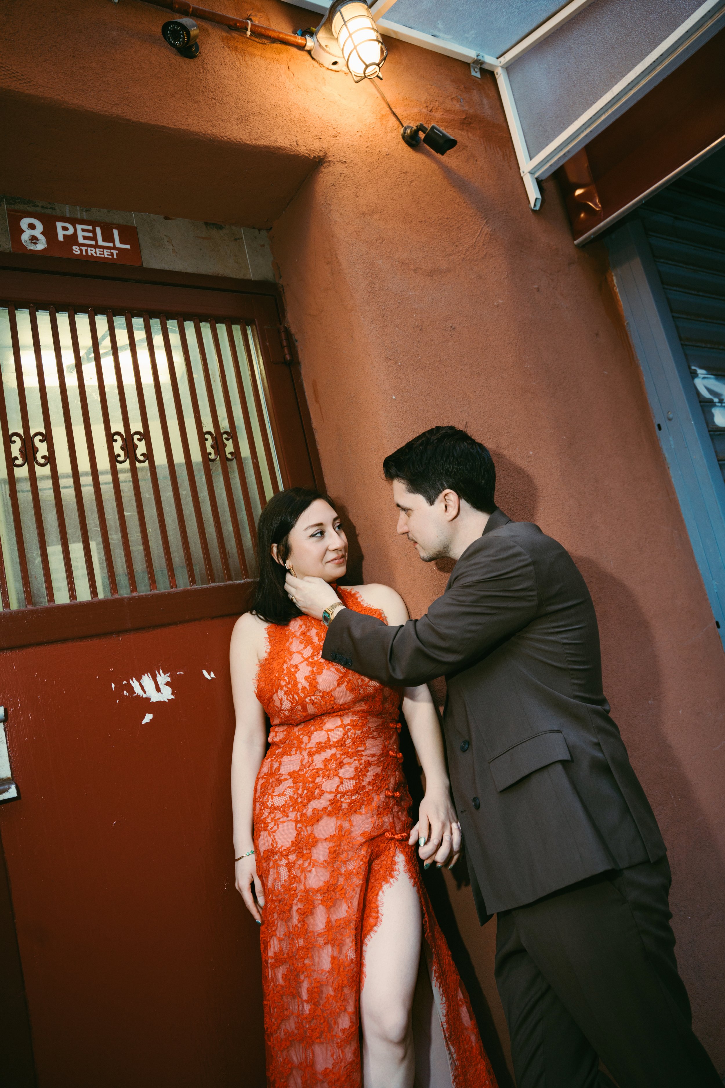 A man and woman are standing close together in front of a red wall, holding hands, with the man gently touching the woman's face. The woman is wearing an orange lace dress with a high slit, and the man is in a dark suit. There is a sign on the wall t