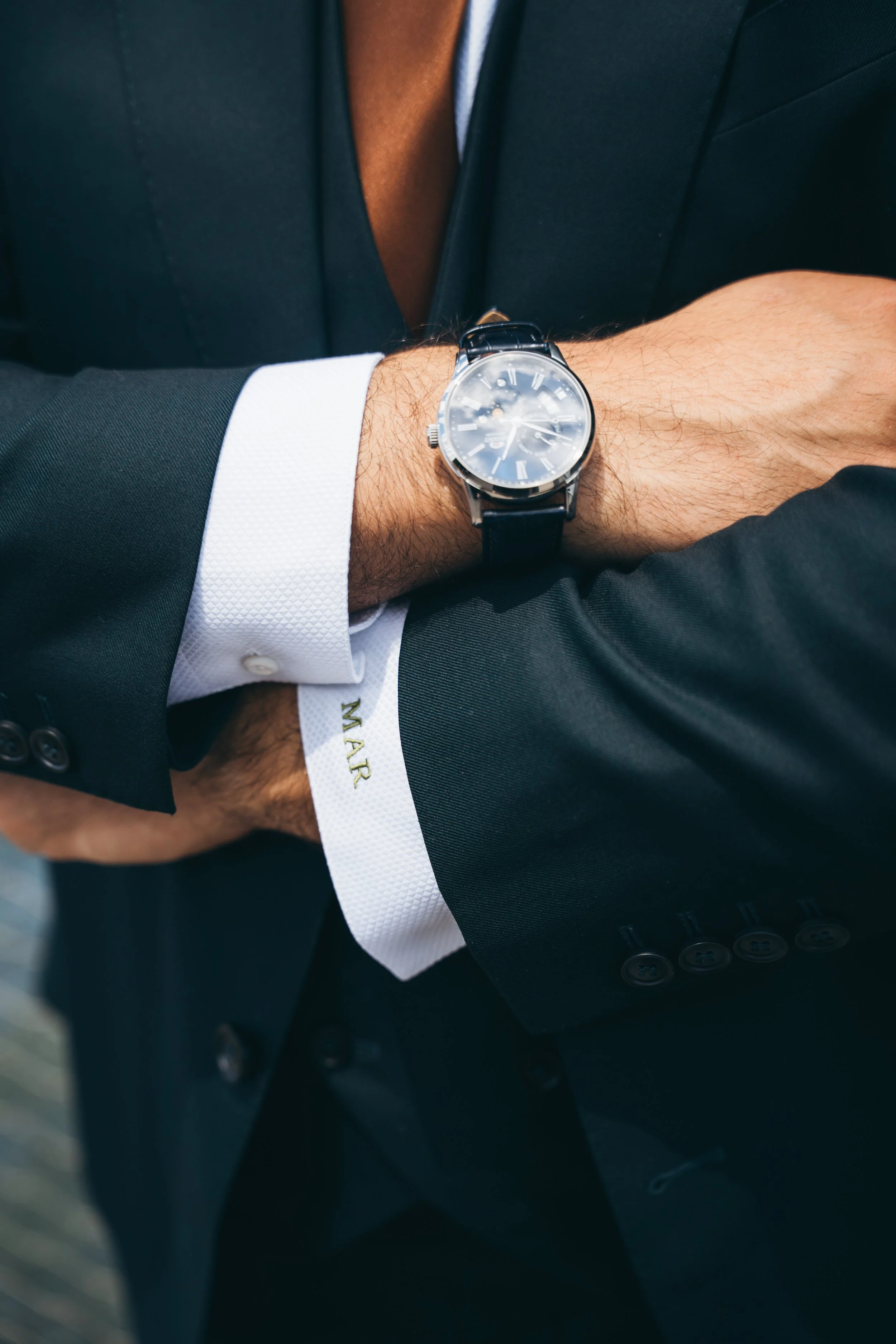 Close-up of a man's wrist with a silver wristwatch with a black leather band, wearing a black suit, white shirt, and with a white wristband underneath, crossing his arms.