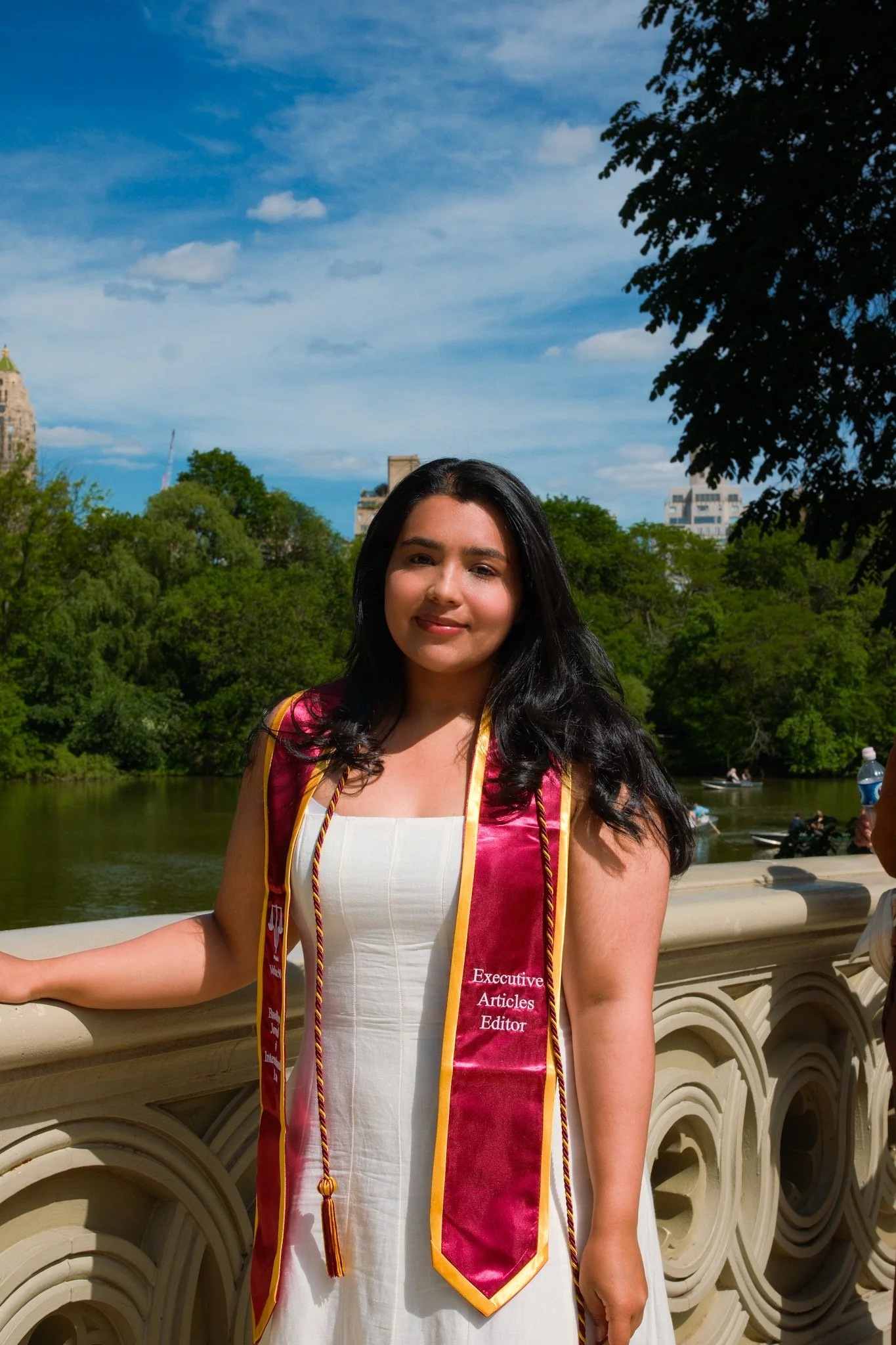 A woman with long black hair smiling, wearing a white dress and a maroon academic stole with gold trim, standing outdoors on a stone bridge with green trees and buildings in the background, under blue sky with some clouds.