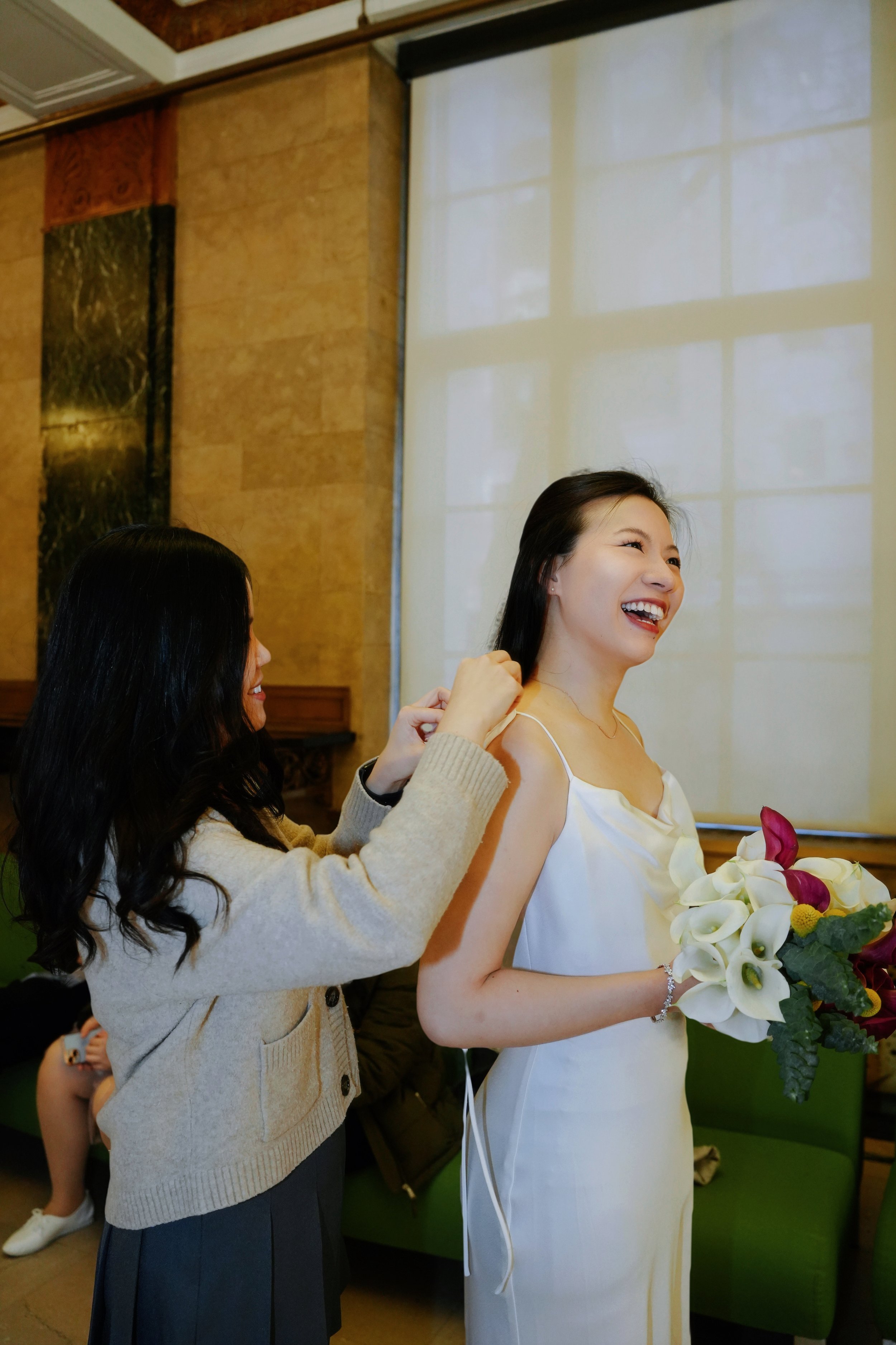 A woman in a satin dress holding a bouquet of white flowers with purple and yellow accents, smiling as another woman pins a piece of jewelry on her.