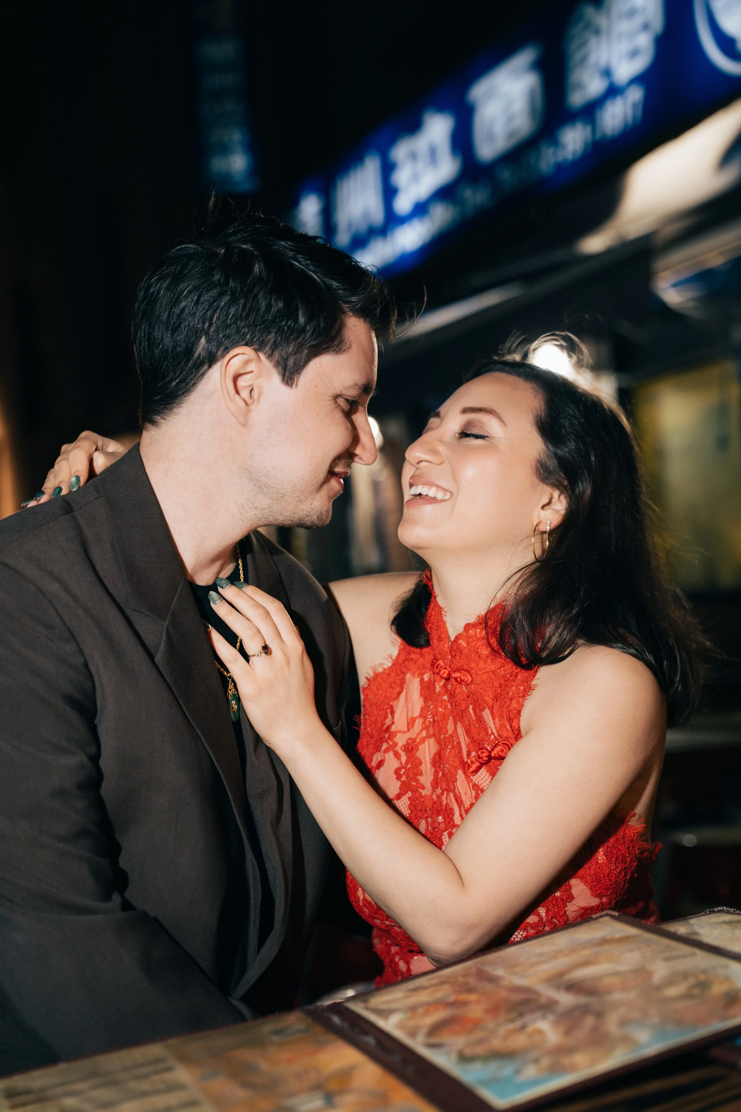 A man and woman are close together, smiling and touching each other's faces in a romantic moment, with a blurry background and a dark setting.