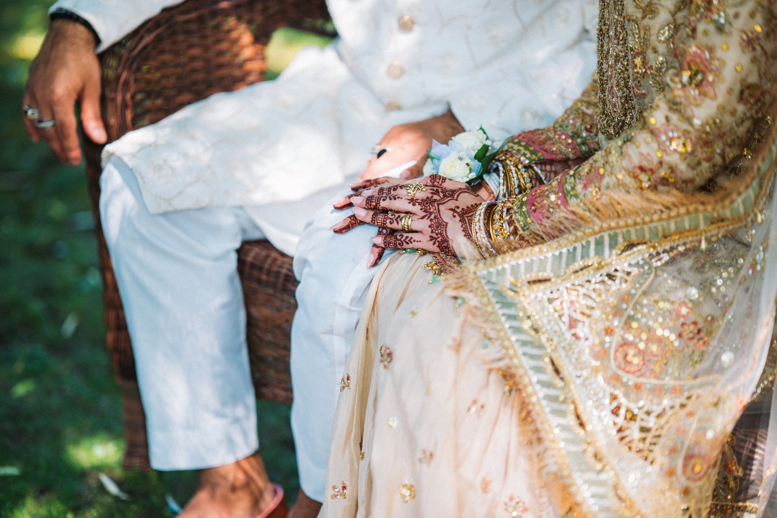 A close-up of a wedding ceremony showing a bride with henna-adorned hands and colorful jewelry, holding a small bouquet of white flowers, sitting on a decorative seat, with part of a groom’s arm and hand visible in the background.