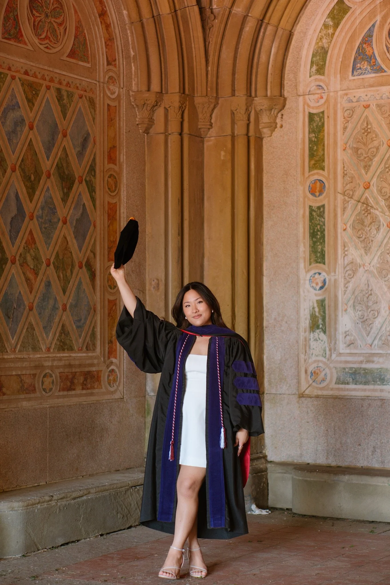 A woman in a graduation gown standing in front of an ornate wall with decorative murals, holding a black hat in her right hand, and posing with her left leg slightly bent.