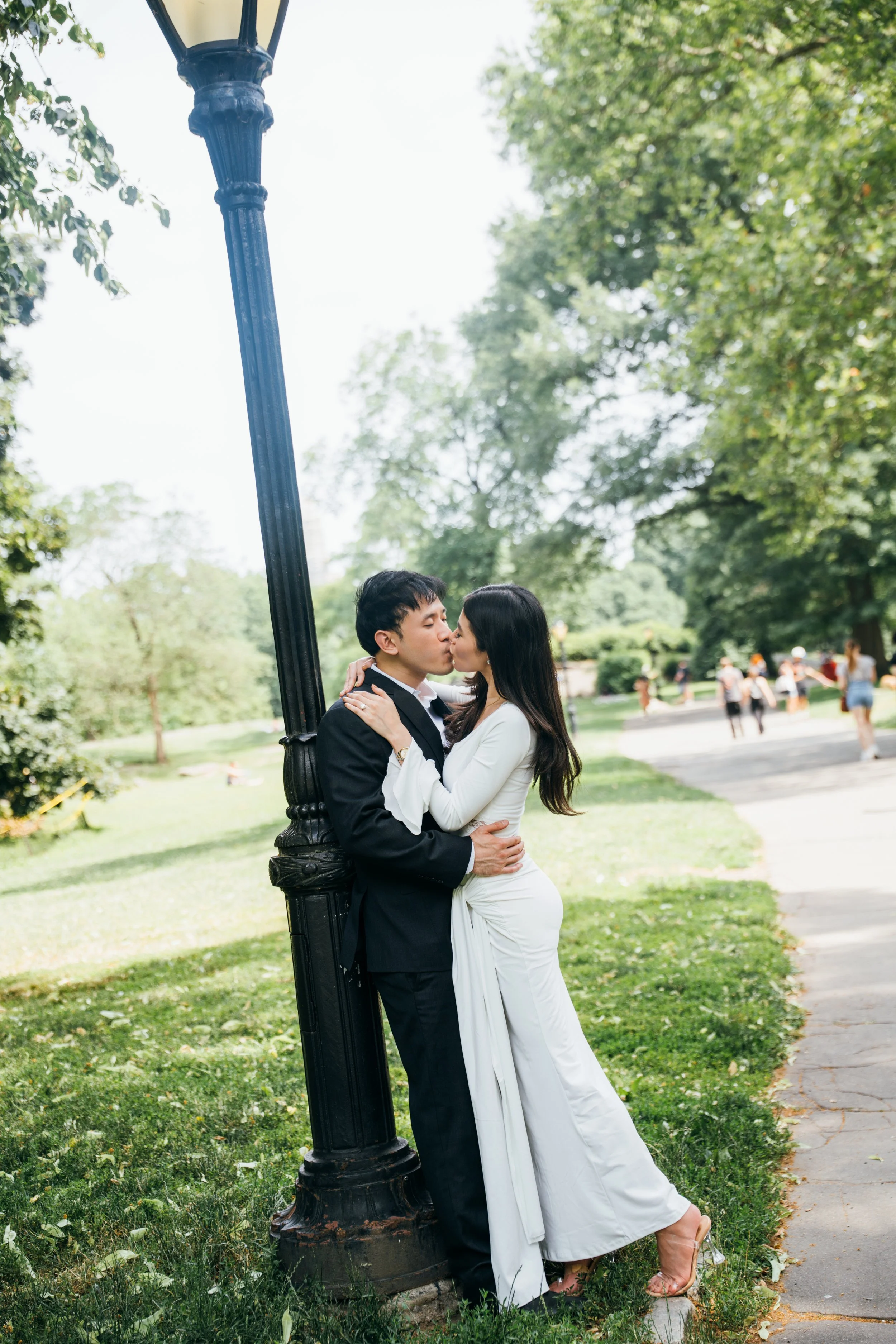 A couple dressed in formal attire sharing a kiss outdoors near a black lamppost in a park with lush green trees and other people walking in the background.