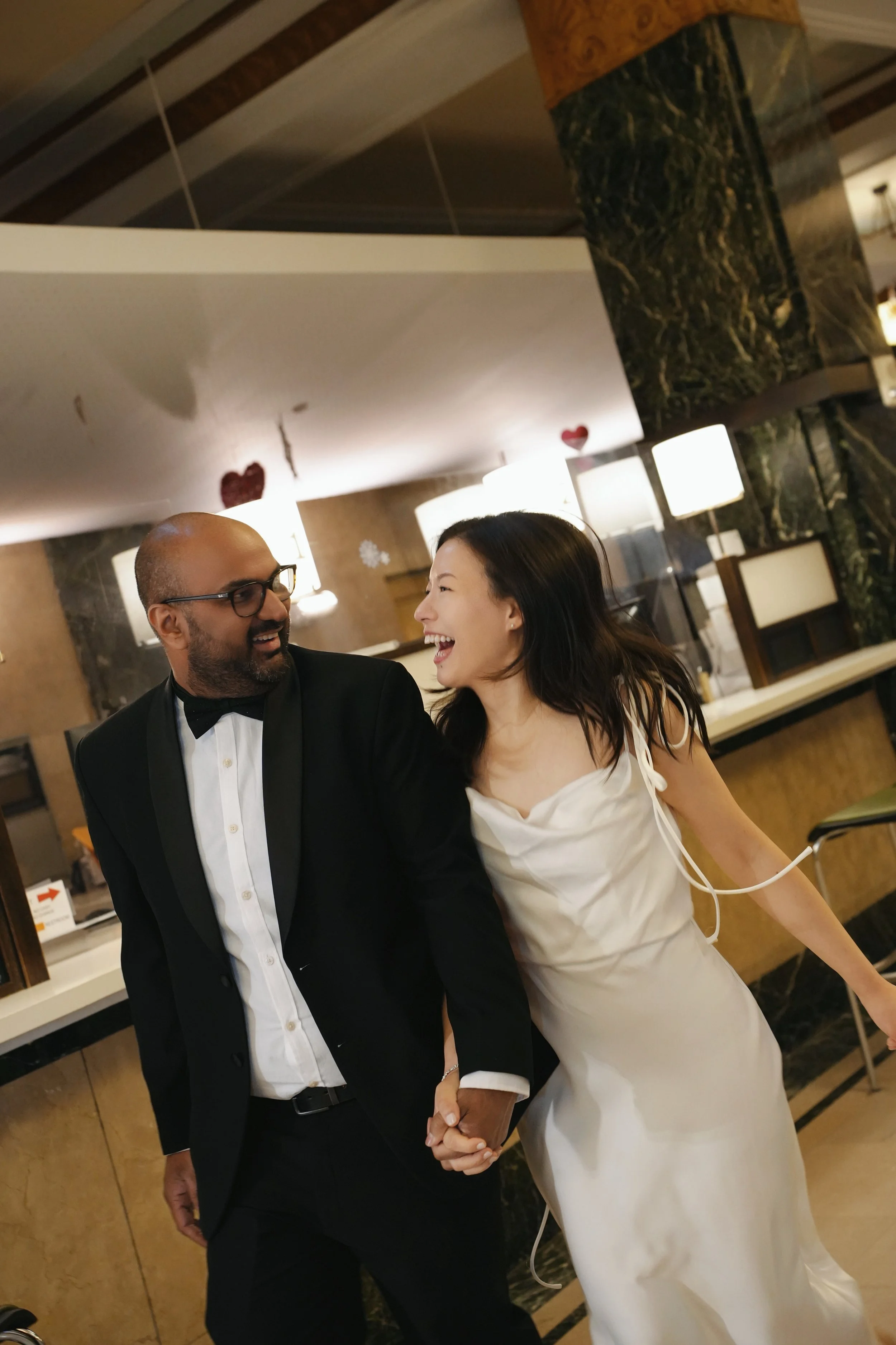 A smiling couple holding hands, dressed in formal attire, attending a special event in an elegant venue decorated with hearts and snowflakes.