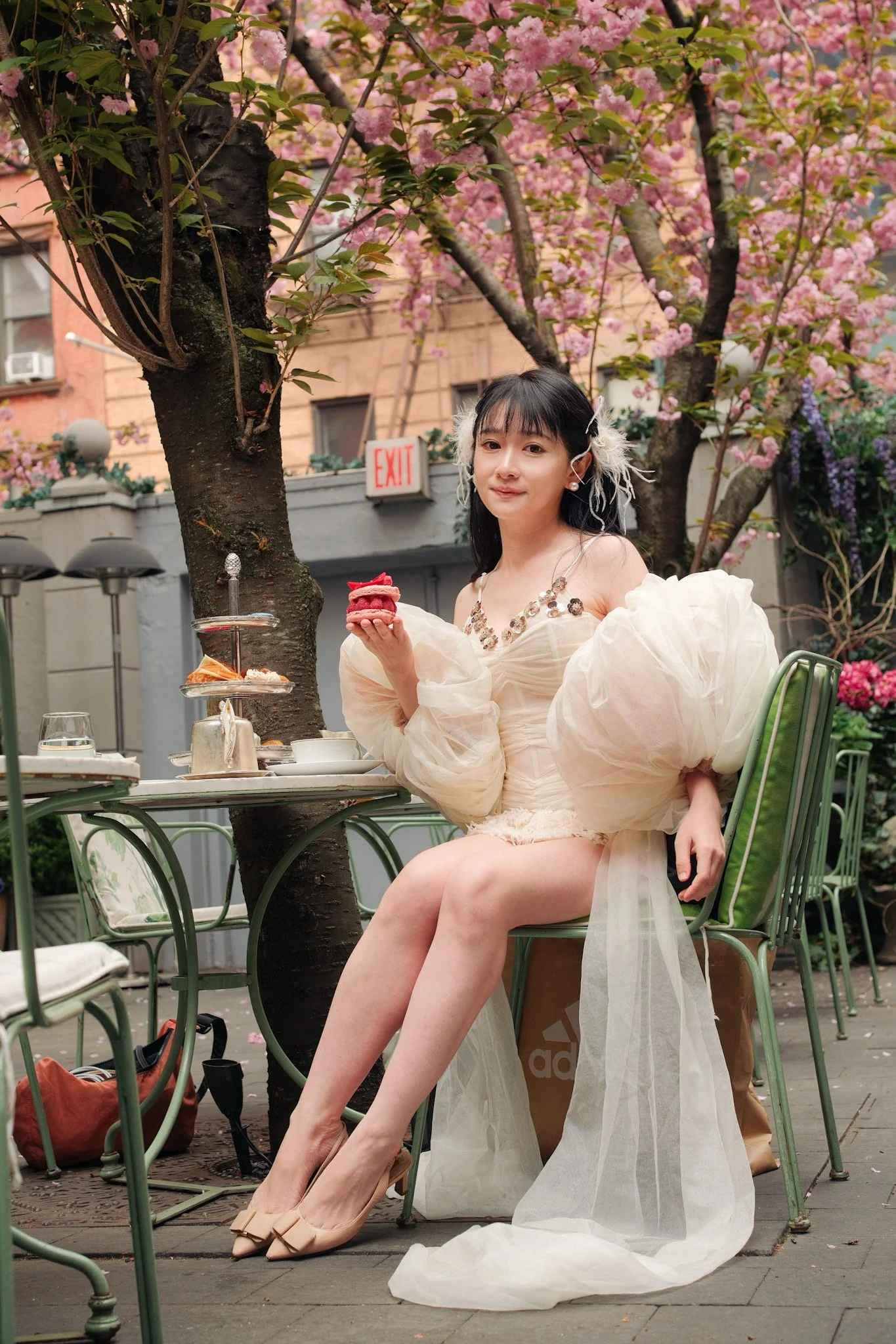 Woman sitting at an outdoor table under pink flowering trees, holding a small dessert and wearing a elegant dress and high heels.