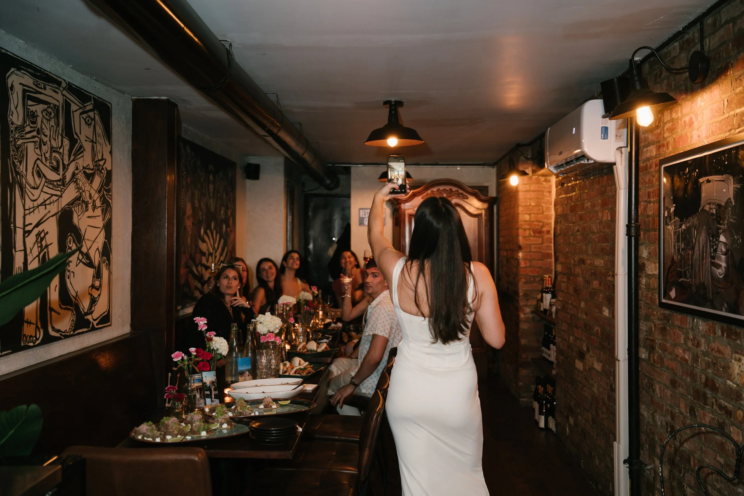 Group of people sitting at a long dining table in a cozy, dimly lit restaurant with brick walls, art, and floral centerpieces, as a woman in a white dress takes a photo.