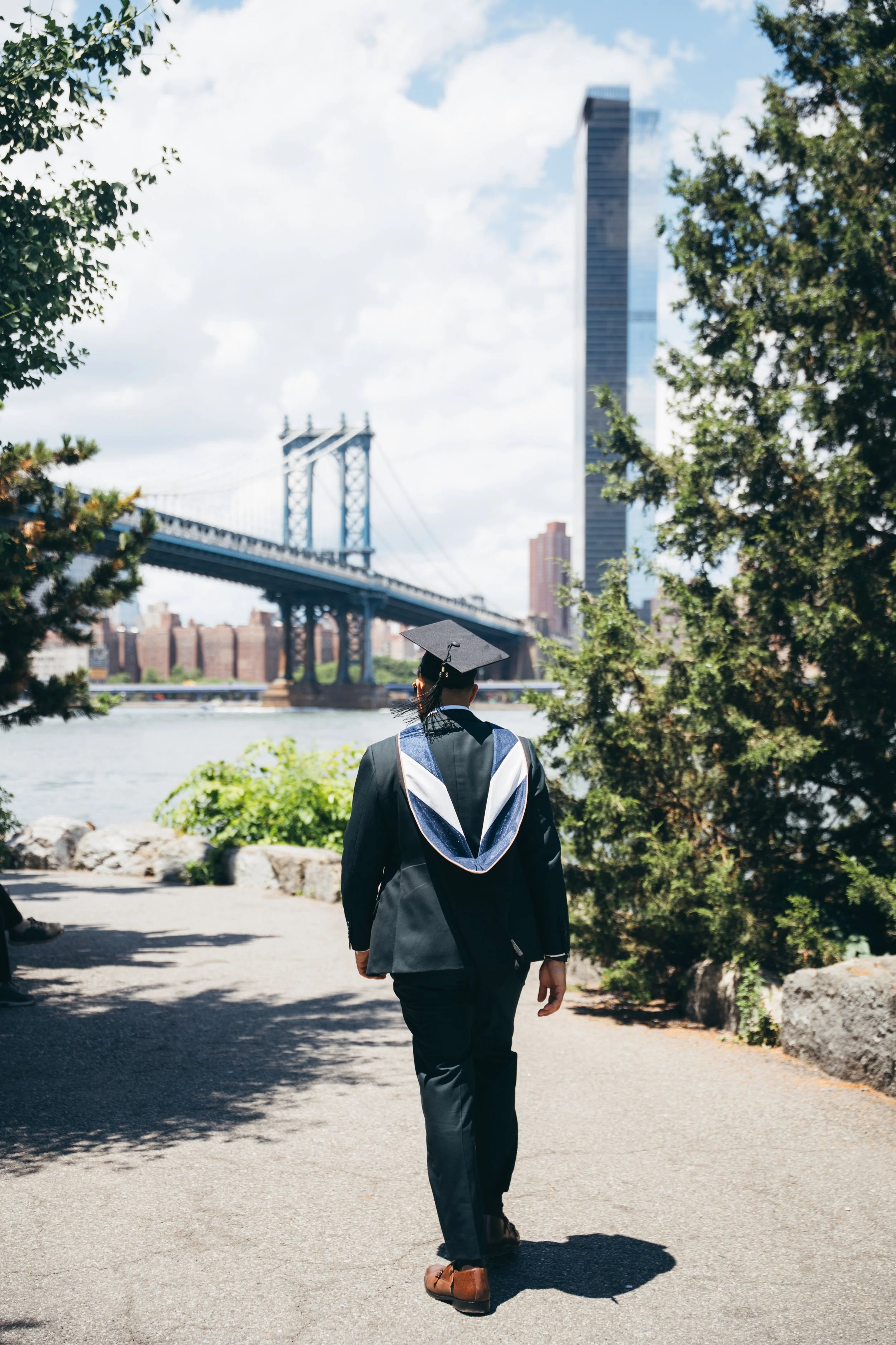 A person dressed in a graduation cap and gown walking on a path next to a river, with trees and tall buildings, including one under construction, in the background.
