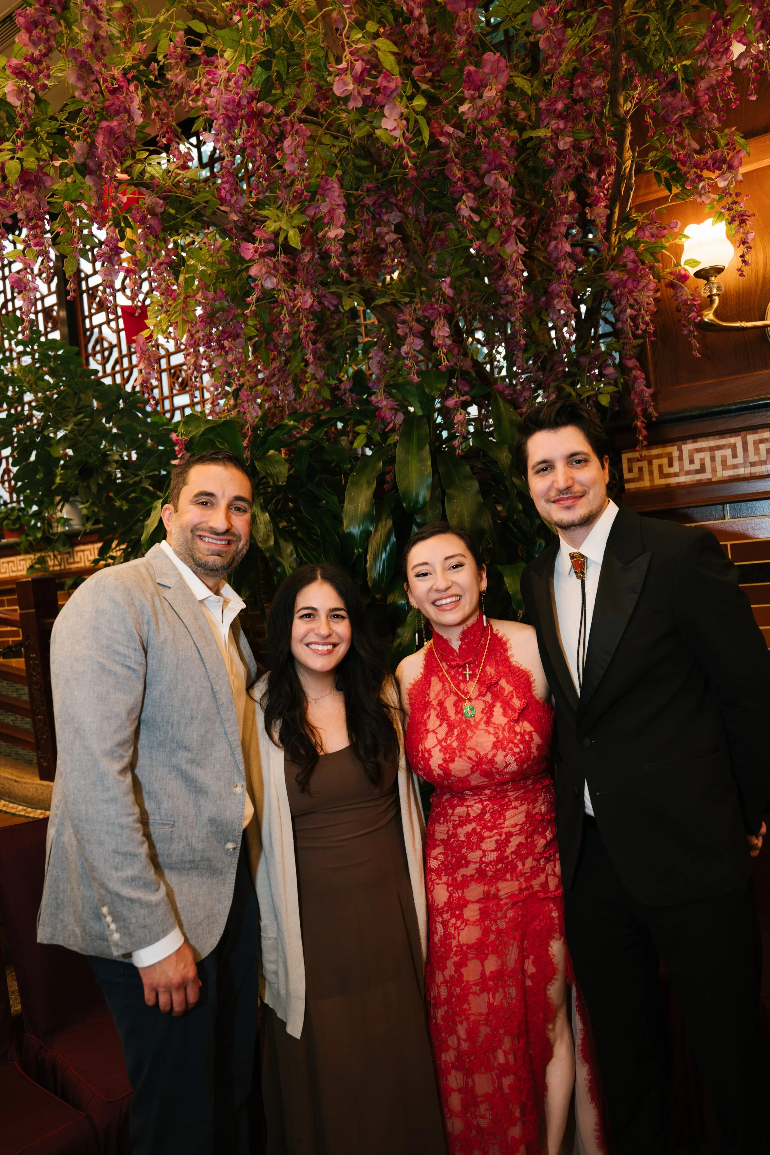Five people, three women and two men, pose together inside a restaurant with a decorative wooden lattice, plants, and a wall sconce. They are dressed formally, smiling at the camera.