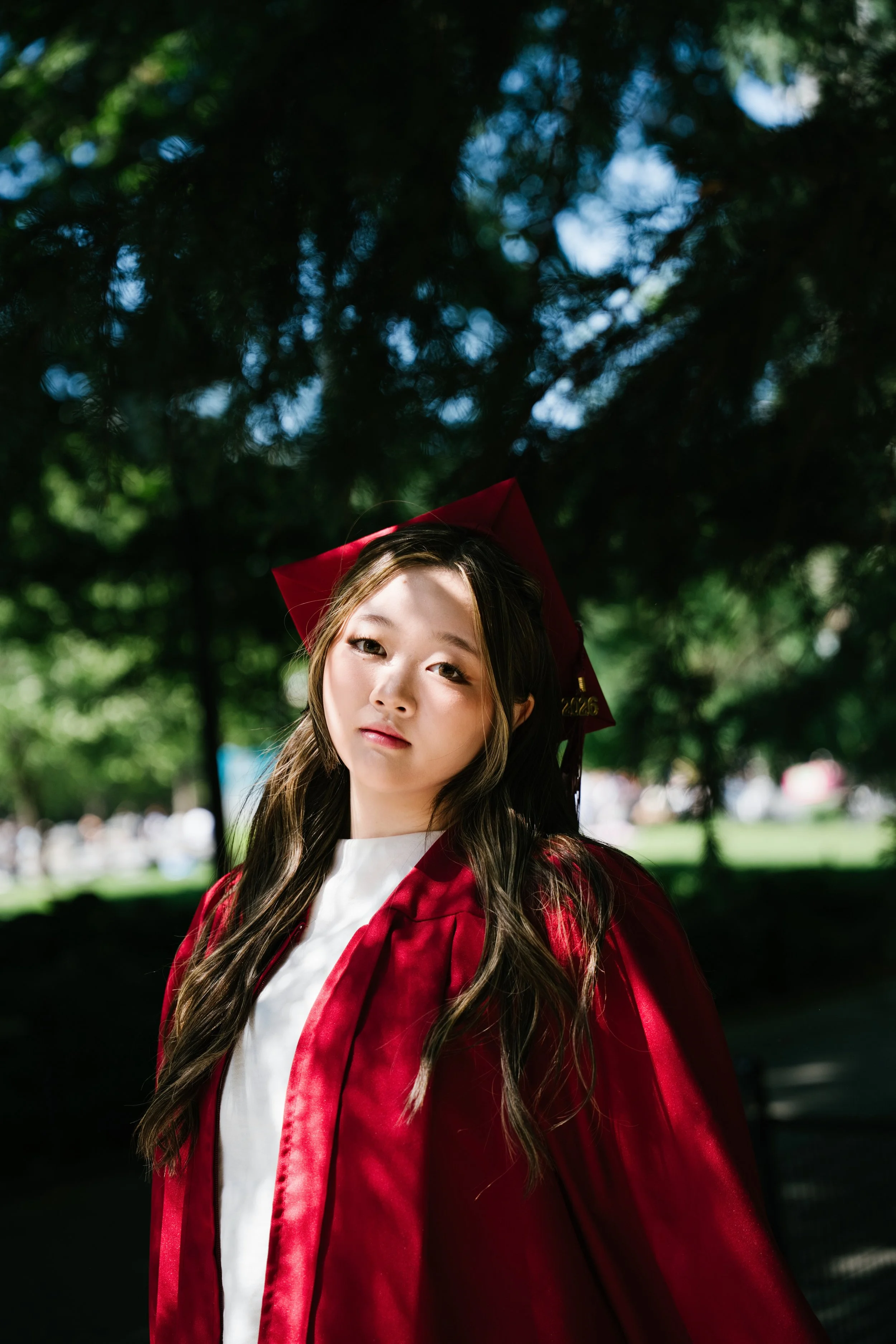 A young woman with long wavy hair wearing a red graduation gown and cap standing outdoors with blurred trees in the background.