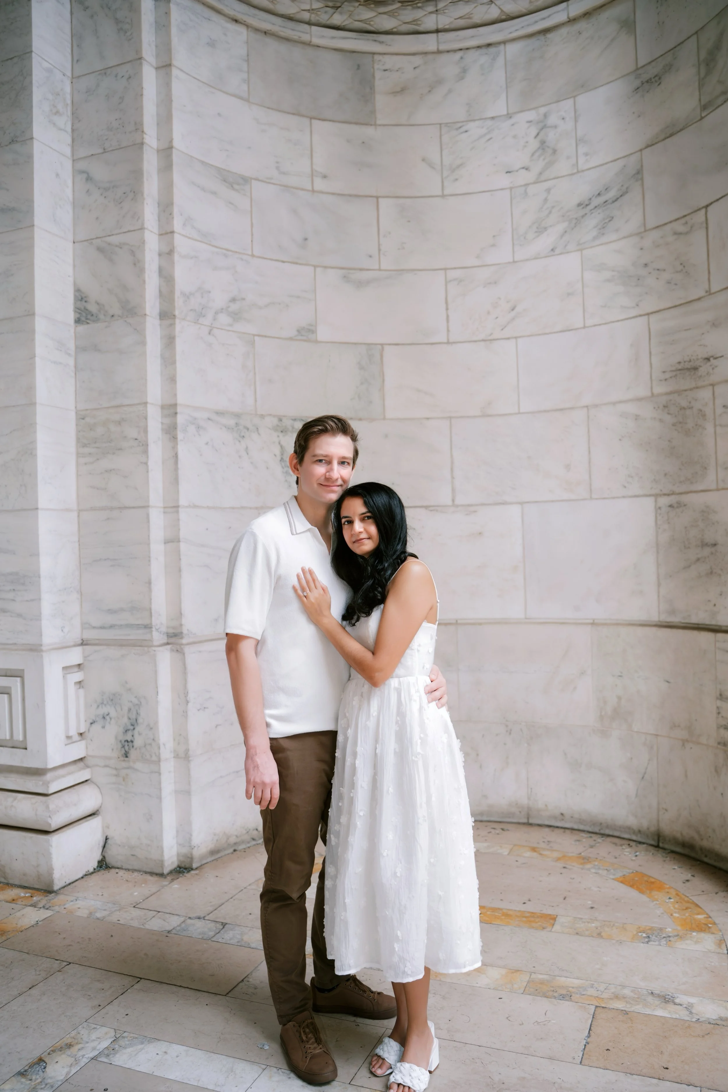A young couple standing close together in front of a large, curved white marble wall. The man has light skin, light brown hair, and is wearing a white shirt and brown pants. The woman has light skin, long black hair, and is wearing a white dress with floral patterns and white sandals. They are holding each other affectionately.