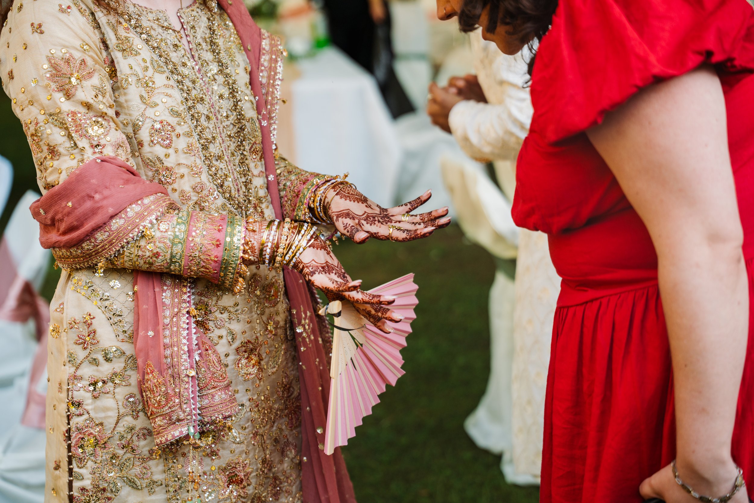 Close-up of traditional Indian wedding attire, showing intricate embroidery, beadwork, and jewelry on the bride's dress, with women in colorful dresses exchanging gifts and flowers at an outdoor celebration.