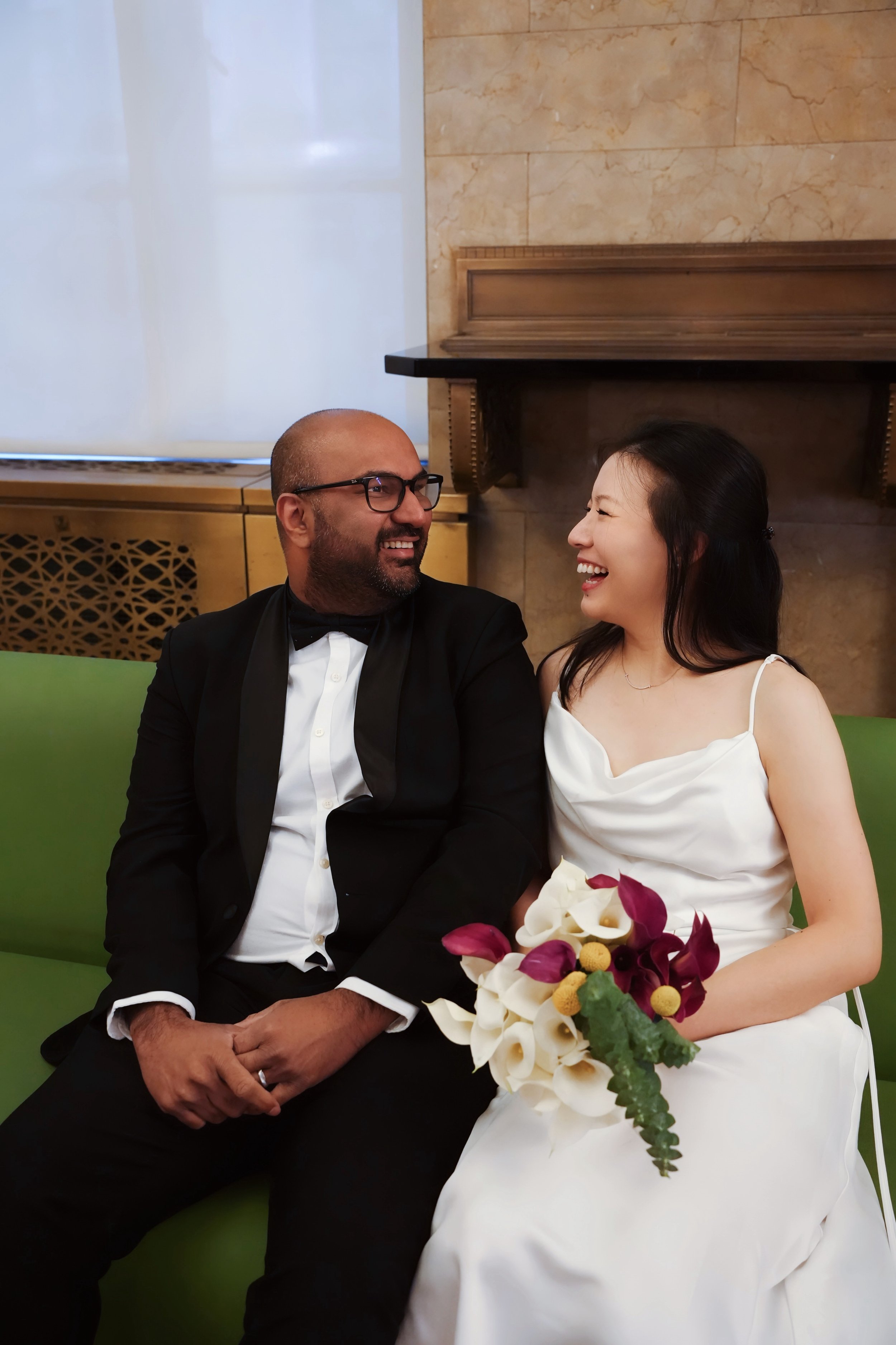 A man in a tuxedo and a woman in a wedding dress sitting on a green bench, smiling and looking at each other, with a bouquet of flowers in front of the woman, in an indoor setting with a marble wall and a fireplace.