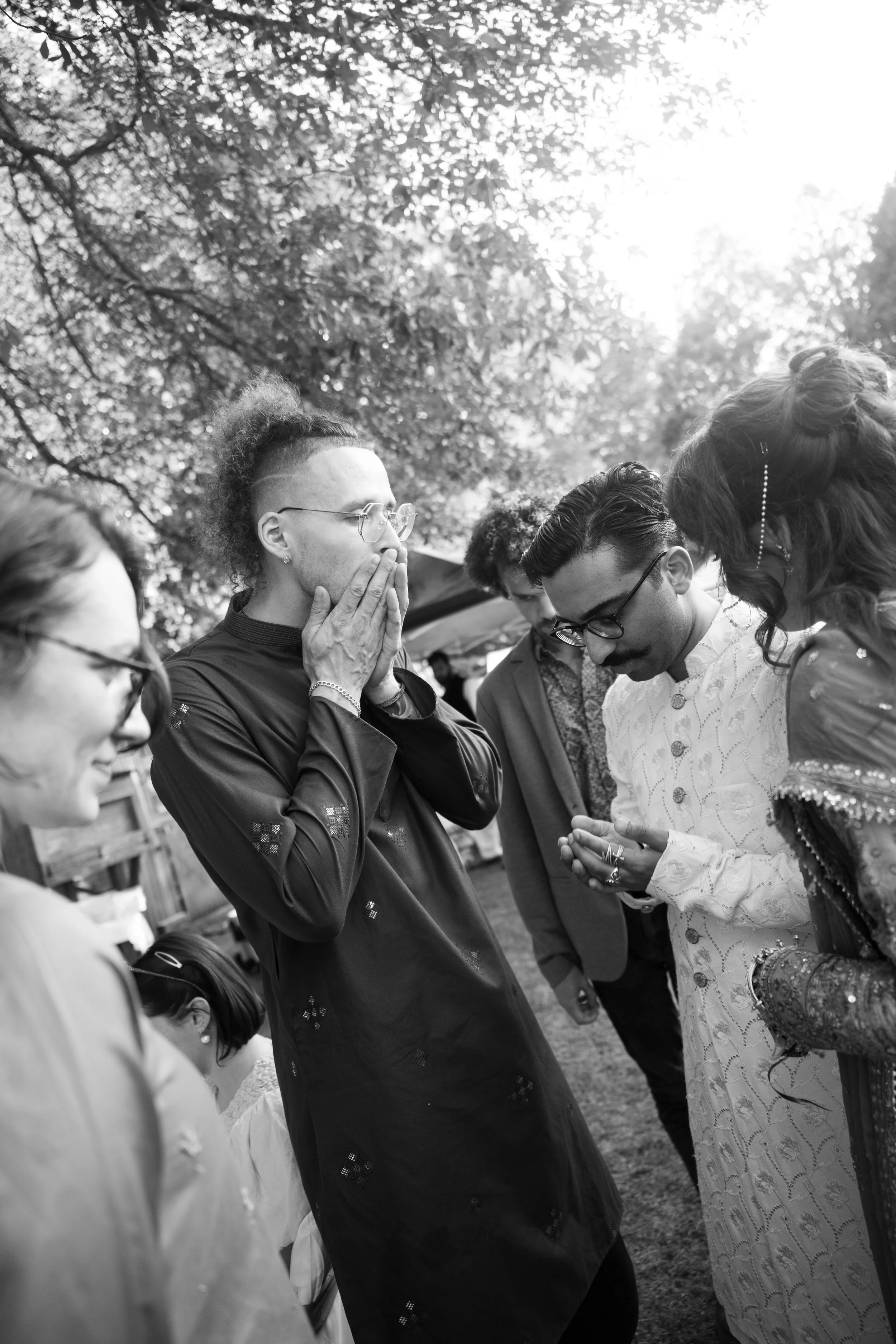 Group of people praying outdoors in a circle, with trees and sunlight overhead.