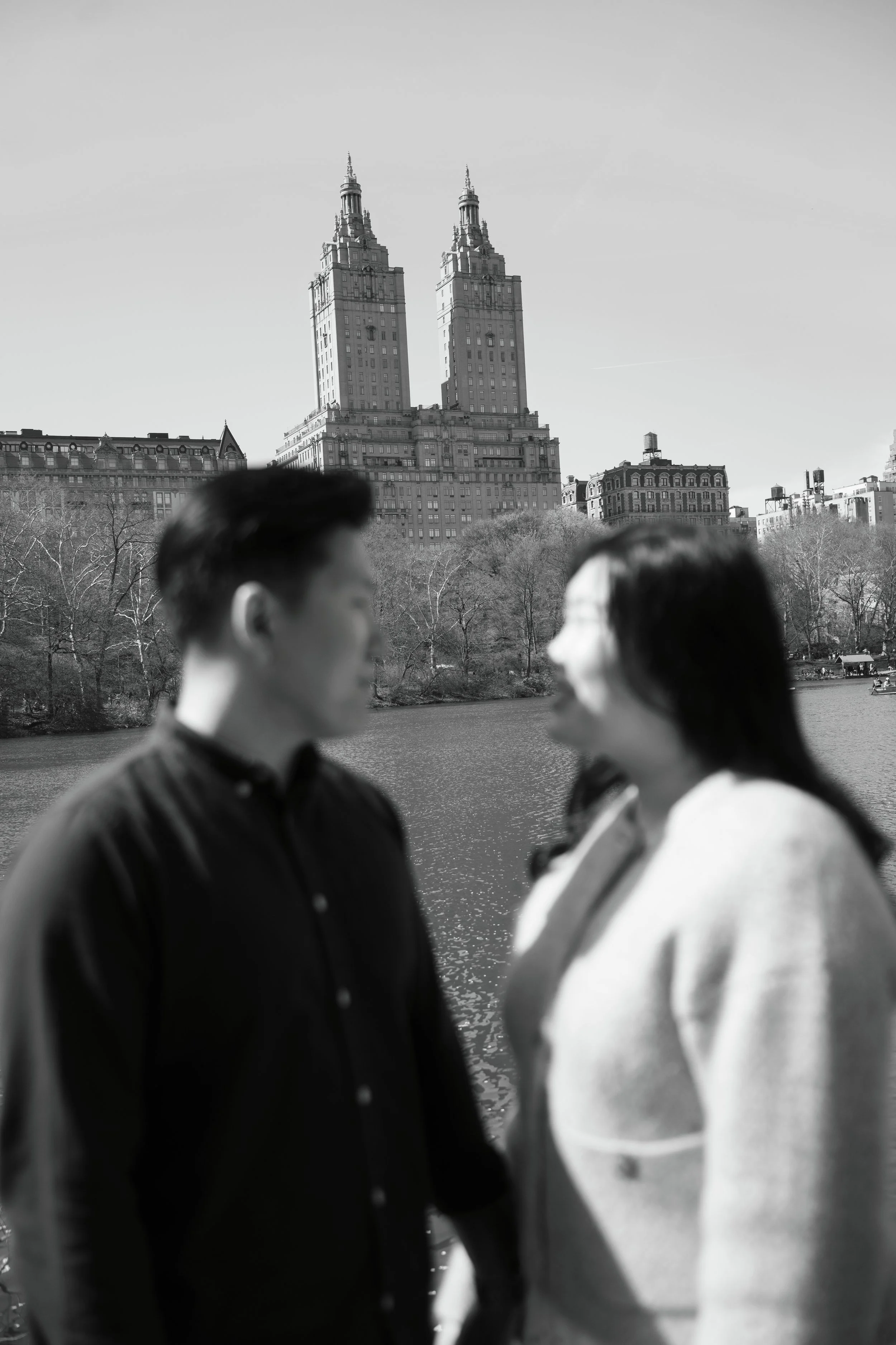 Black and white photo of two people in the foreground, a man and a woman, facing each other near a body of water. In the background, there is a cityscape with tall buildings and trees.