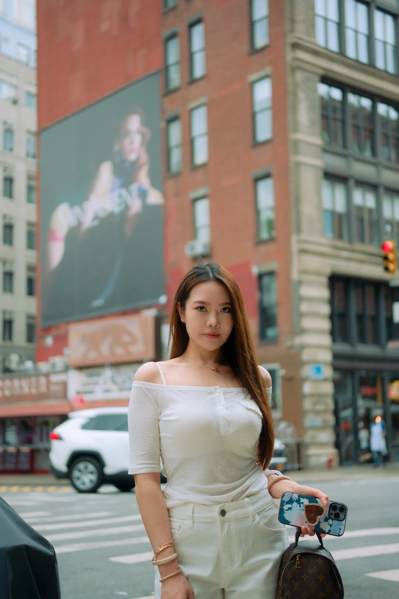 Young woman with long brown hair standing on city street, holding a smartphone and a Louis Vuitton purse, with tall buildings and a large billboard in the background.