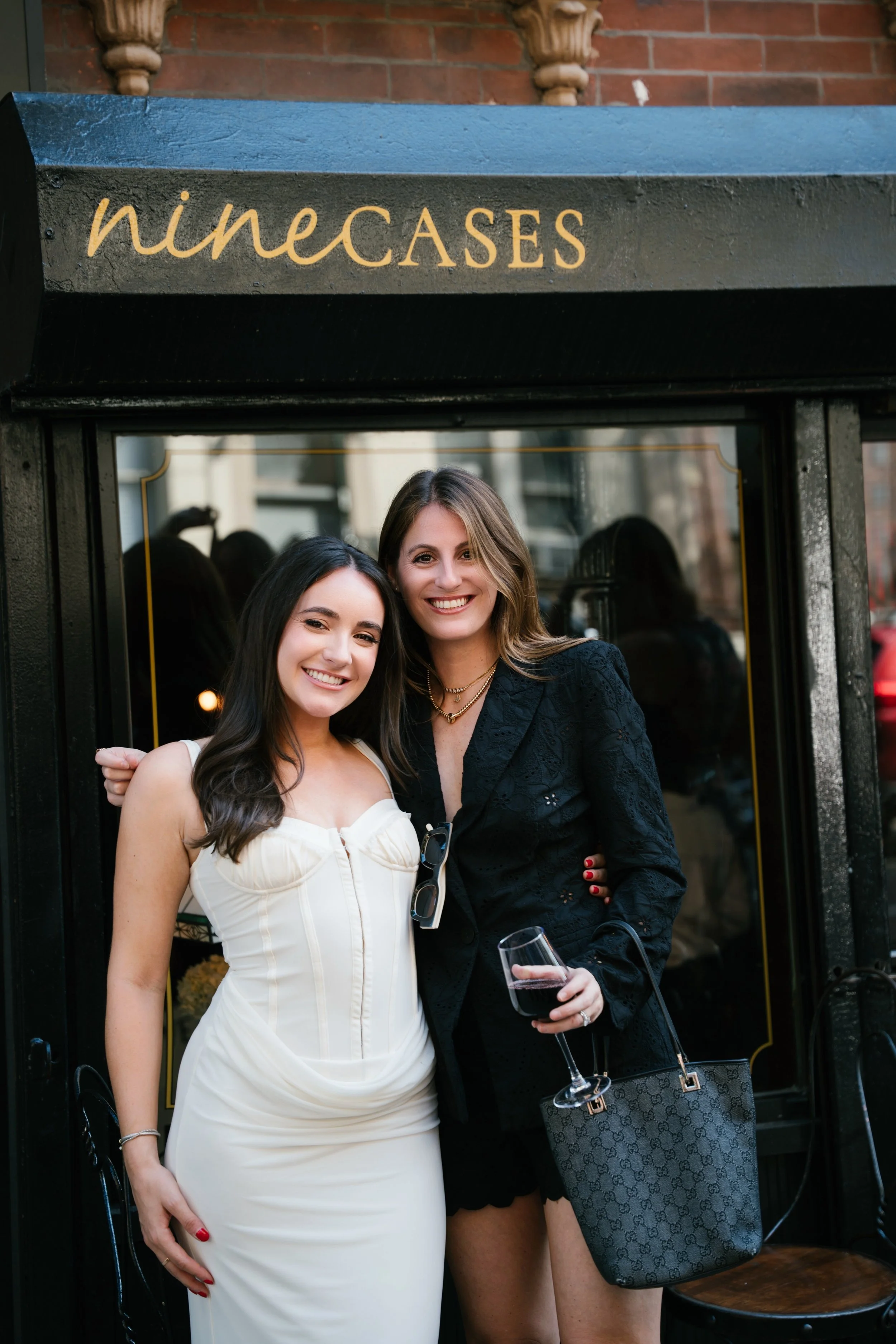 Two women standing together smiling outside near a brick wall and a black storefront with gold lettering that reads 'nineCAGES'.