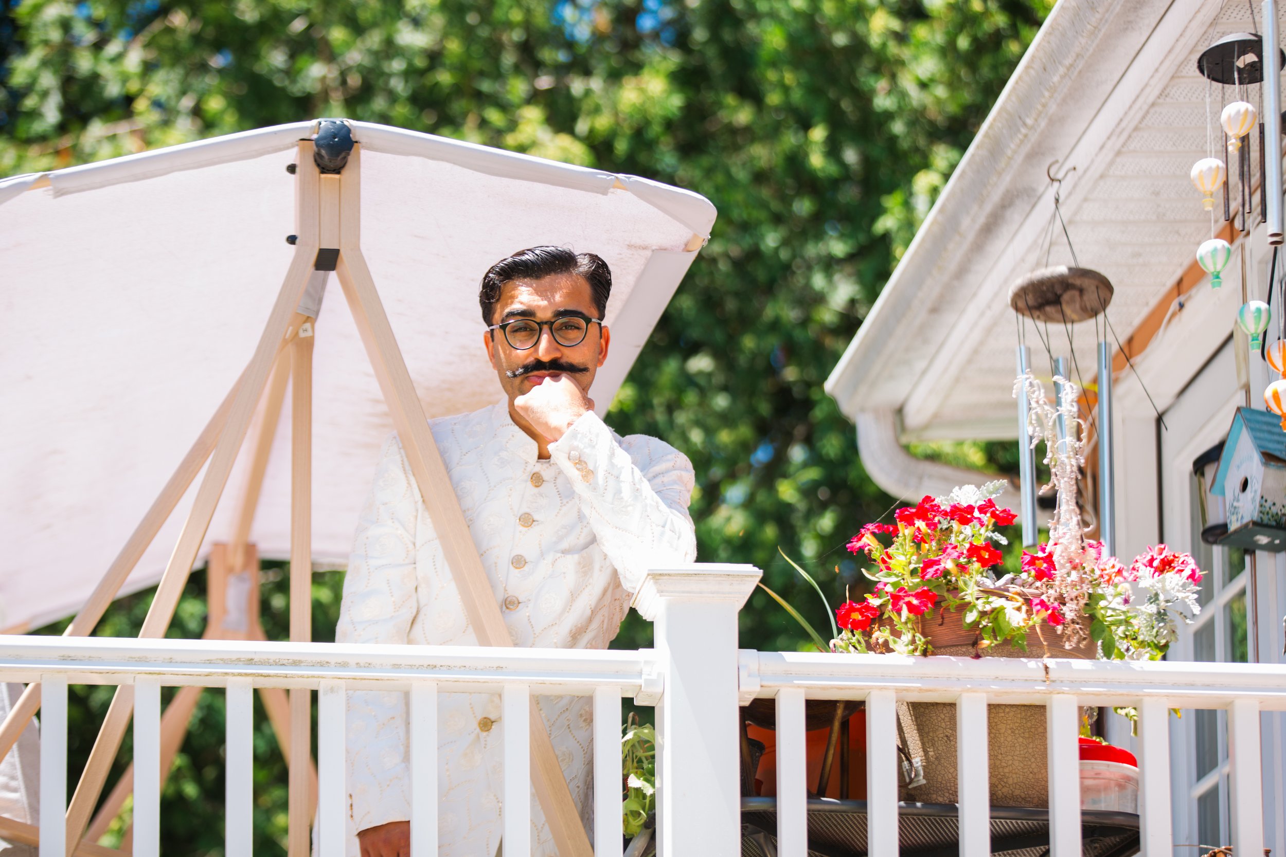A man with glasses and a mustache wearing a traditional white embroidered kurta, standing on a porch or balcony, resting his chin on his hand, surrounded by colorful flowers and outdoor decorations on a sunny day.