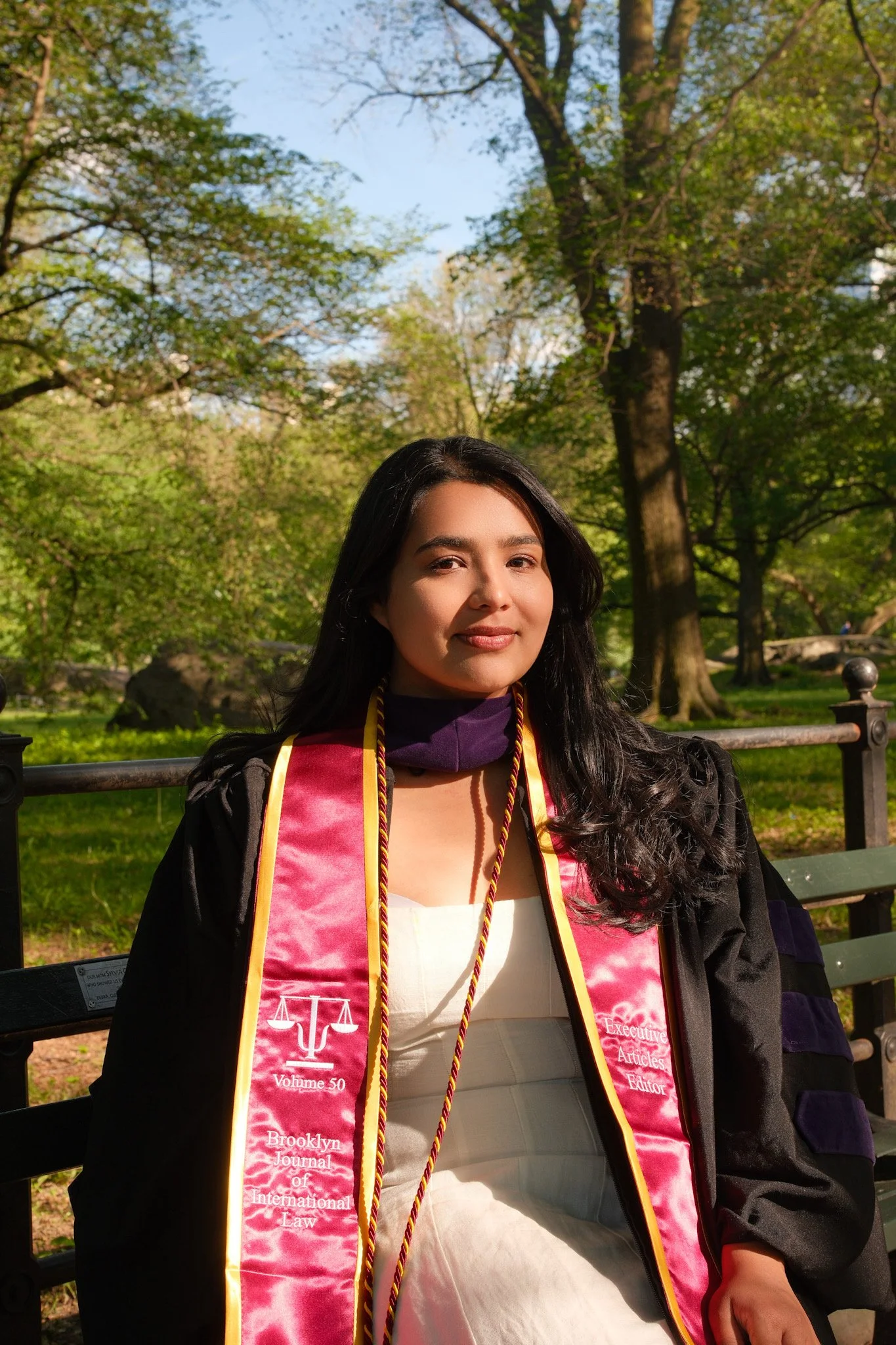 Young woman in graduation gown and pink stole with text, sitting on a park bench in green outdoor setting with trees and blue sky.