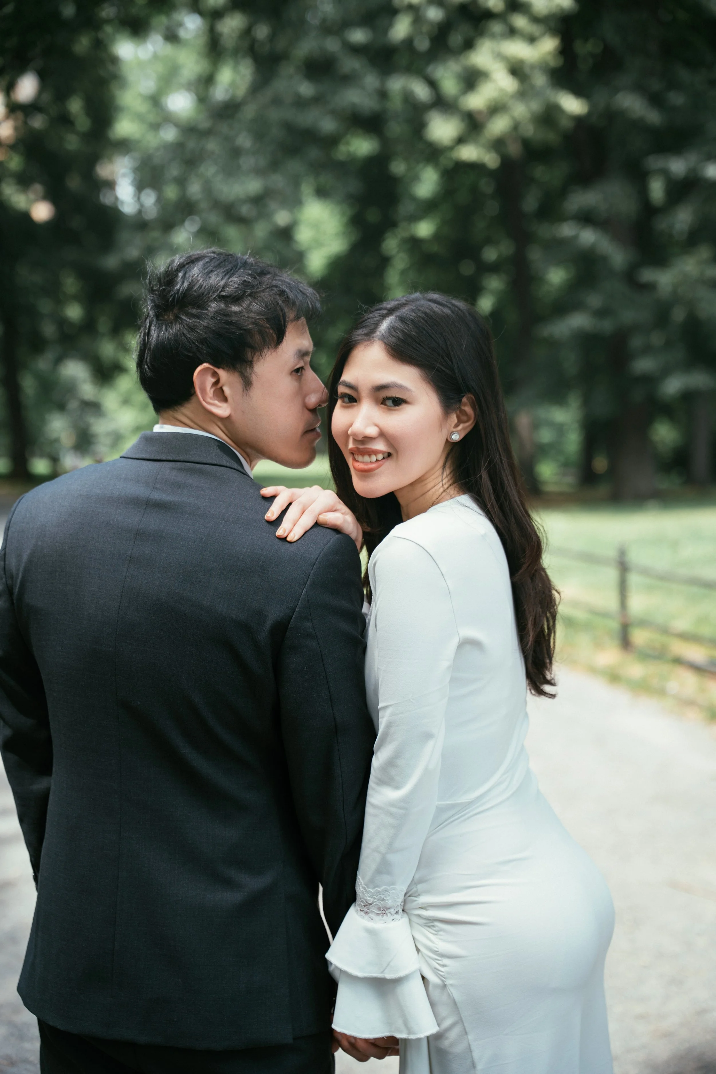 A couple dressed in formal attire, holding hands and standing close together outdoors in a park with green trees in the background.