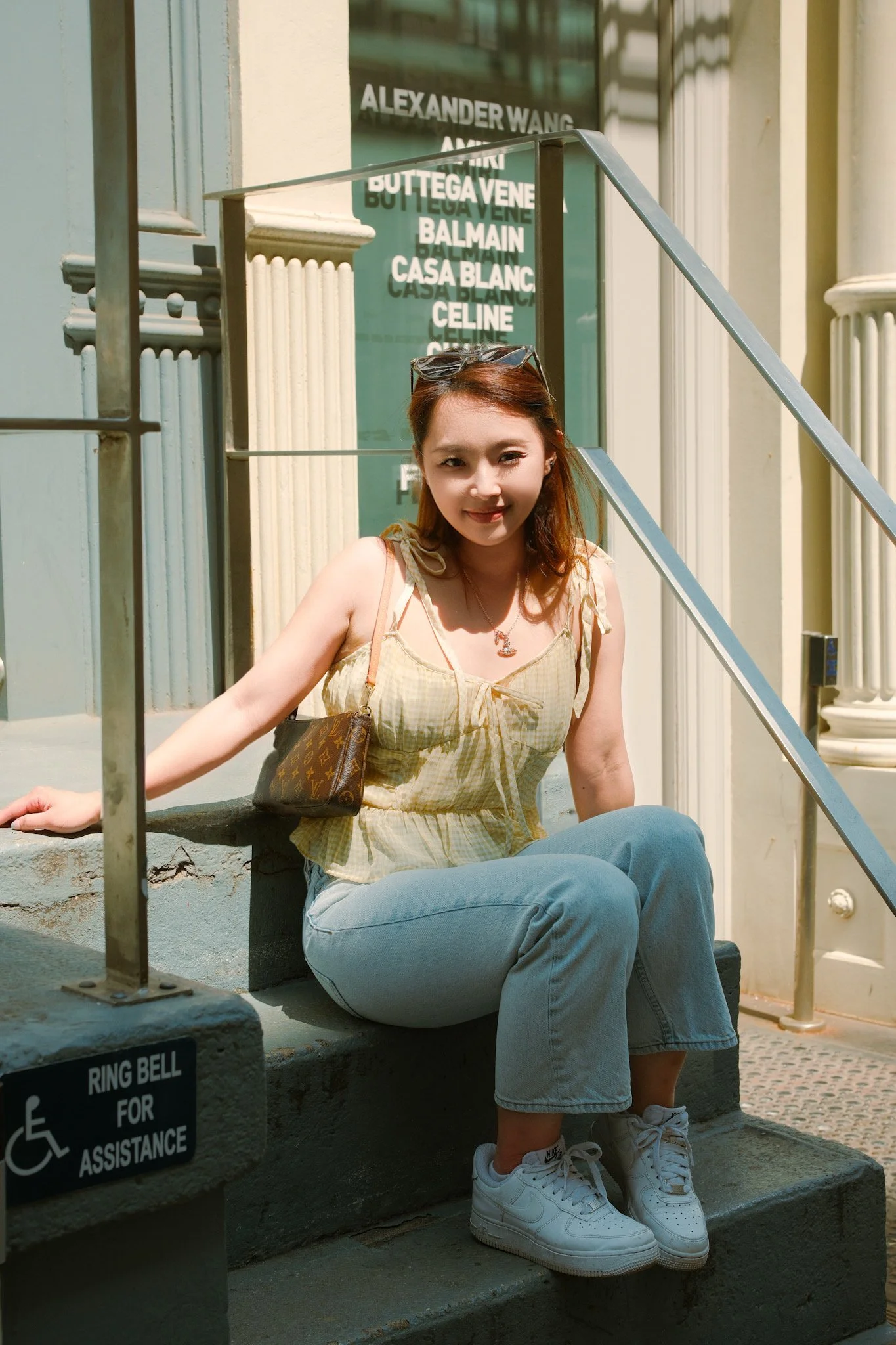 A young woman with reddish-brown hair sitting at the bottom of stairs outside a store, smiling at the camera. She is wearing a yellow plaid sleeveless top, light blue jeans, and white Nike sneakers. A pair of sunglasses rests on her head, and a Louis