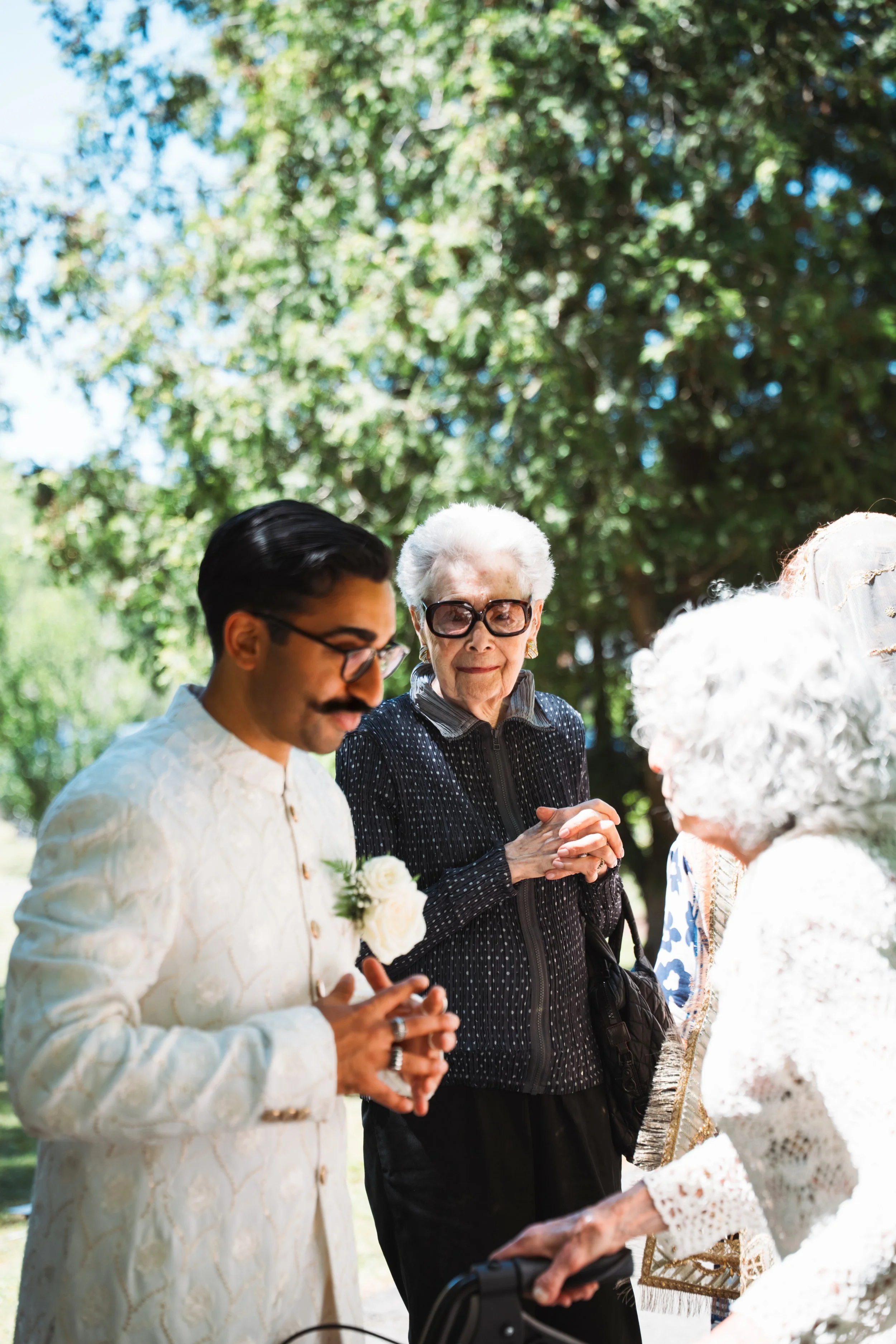 A wedding ceremony outdoors with a bride and groom holding hands and an officiant standing between them, all under a green leafy tree.