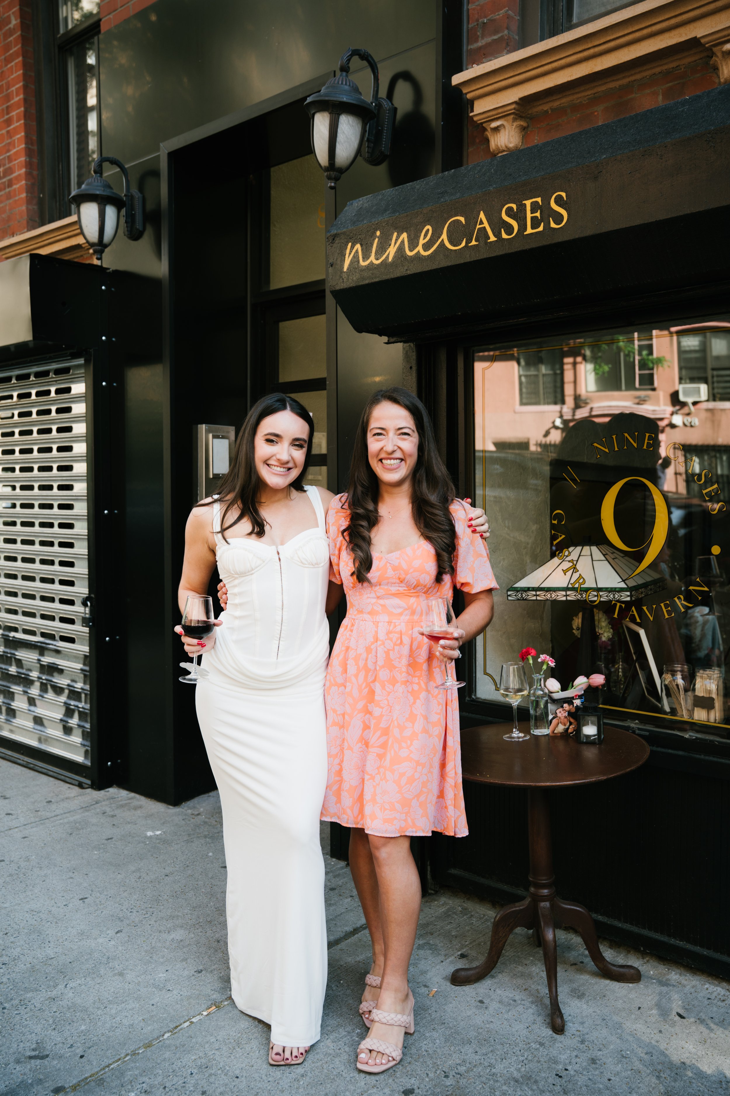 Two women standing outside a restaurant, one in a long white dress holding a glass of red wine, the other in a pink floral dress holding a glass of wine, smiling at the camera.