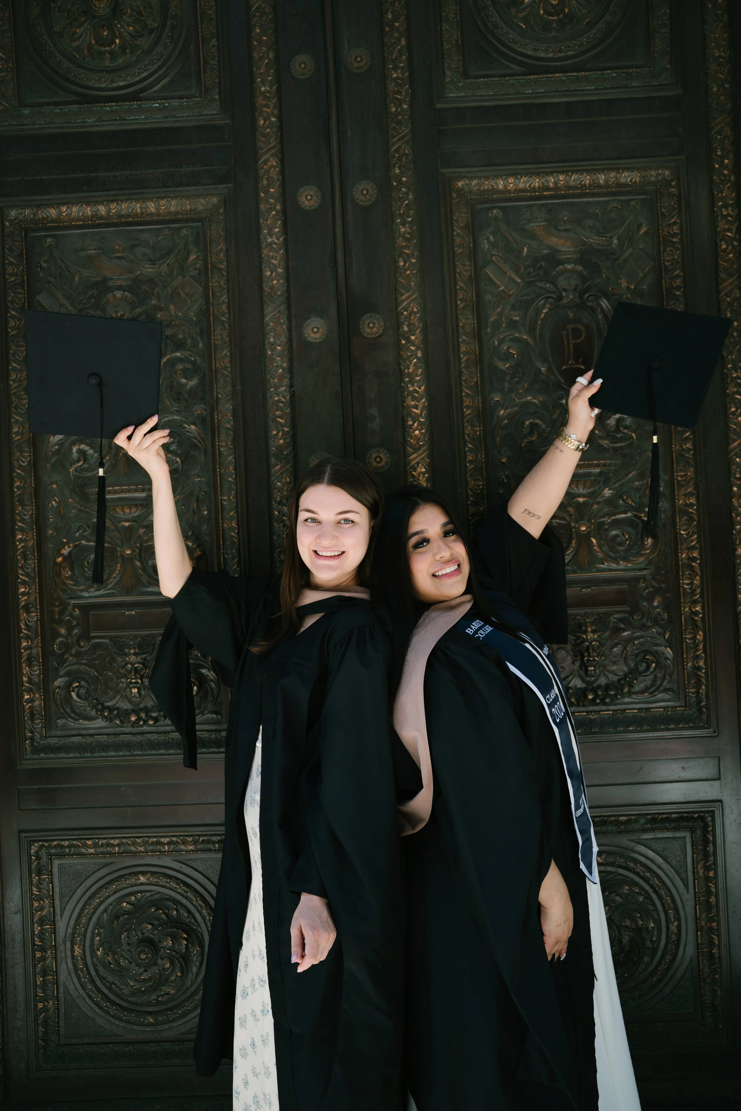 Two young women in graduation robes and caps, holding their caps up, smiling, standing in front of large ornate wooden doors.