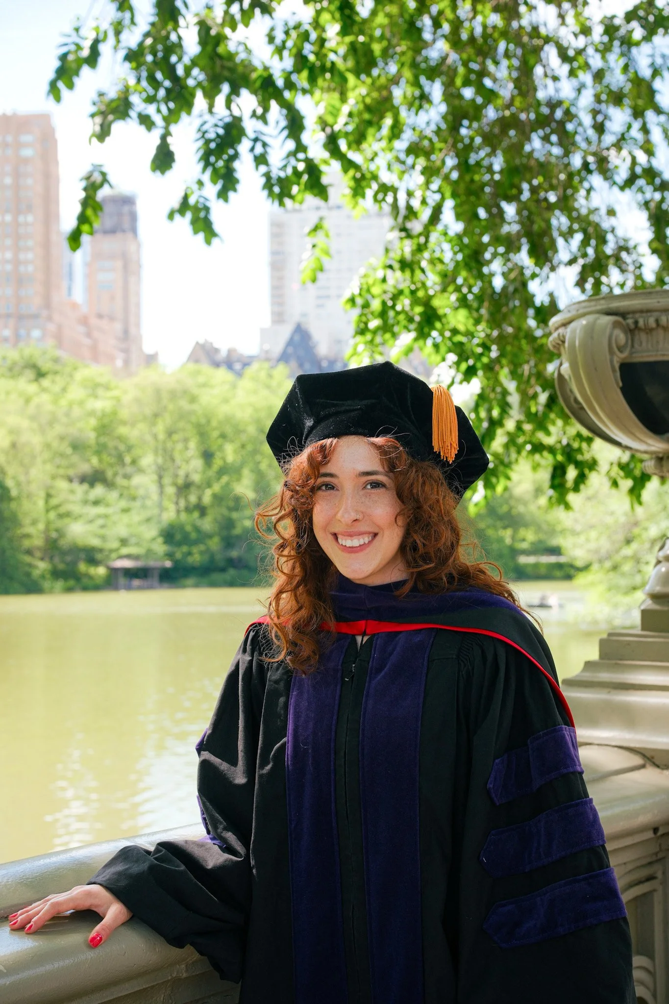A young woman with curly red hair and a big smile is wearing a black graduation cap with an orange tassel and a black and purple gown. She is standing outdoors by a body of water with green trees and tall buildings in the background, under a bright b
