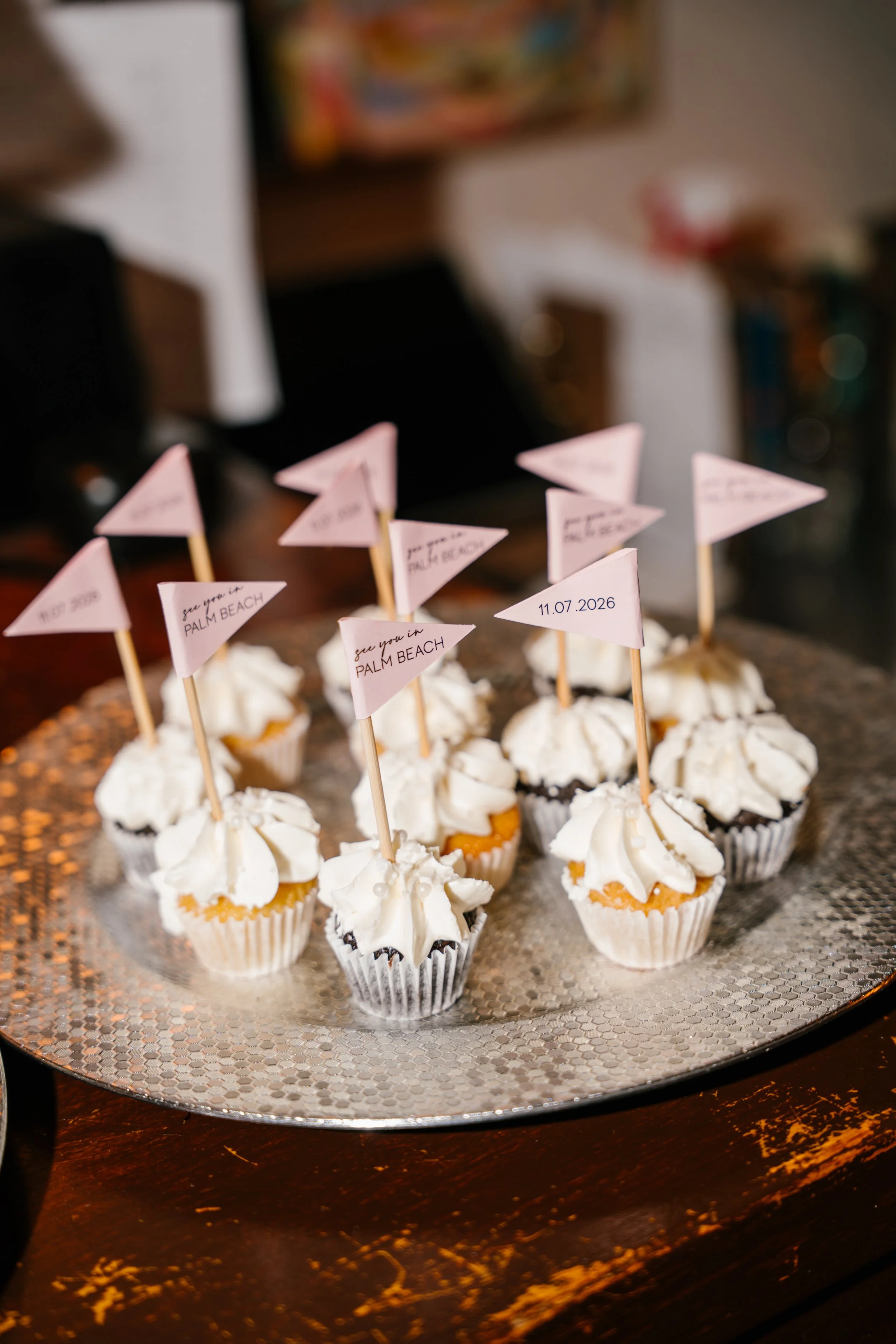 Cupcakes topped with whipped cream and small pink flags with writing, placed on a round silver tray.